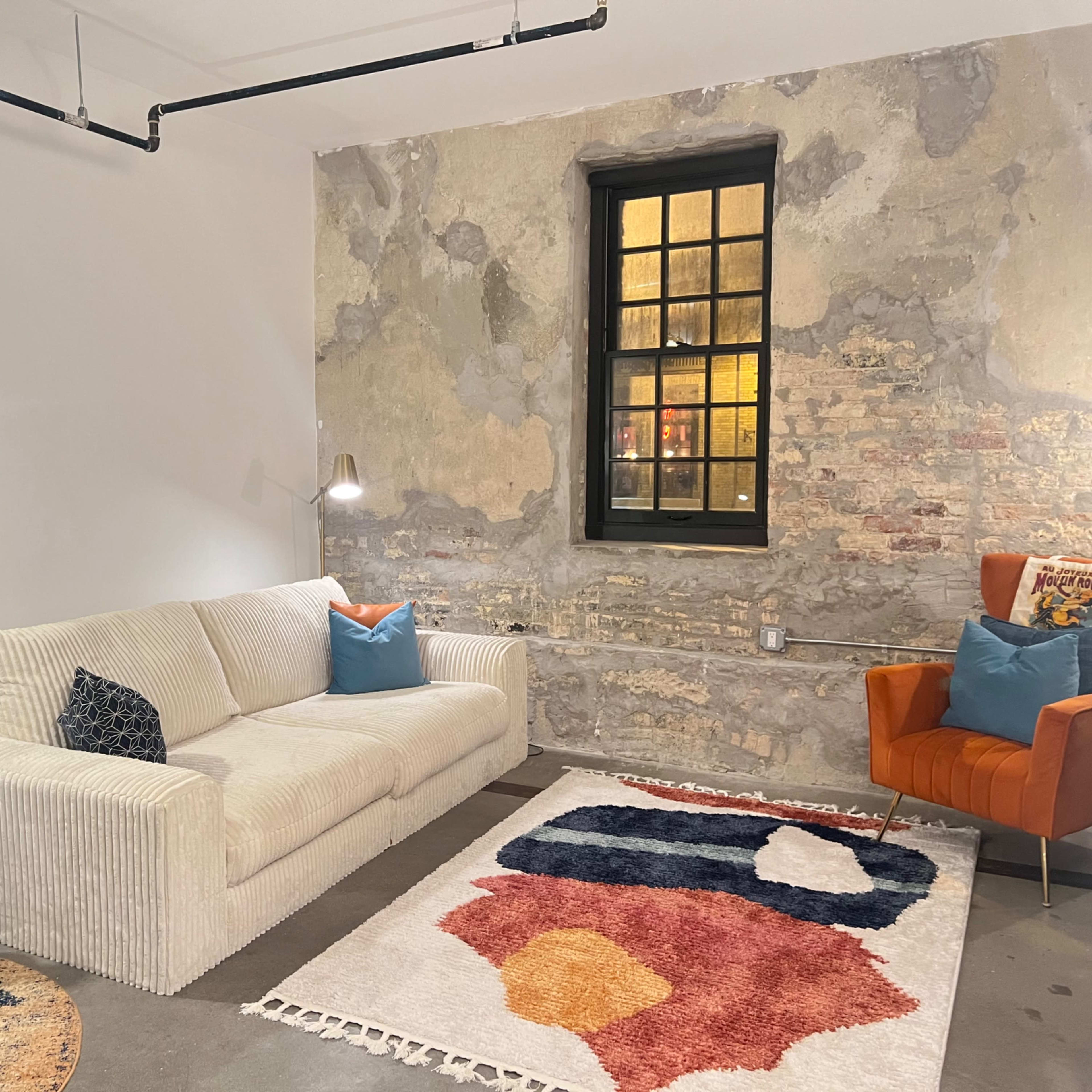 A living room features a white sofa, an orange armchair, a decorative rug, and exposed brick walls.