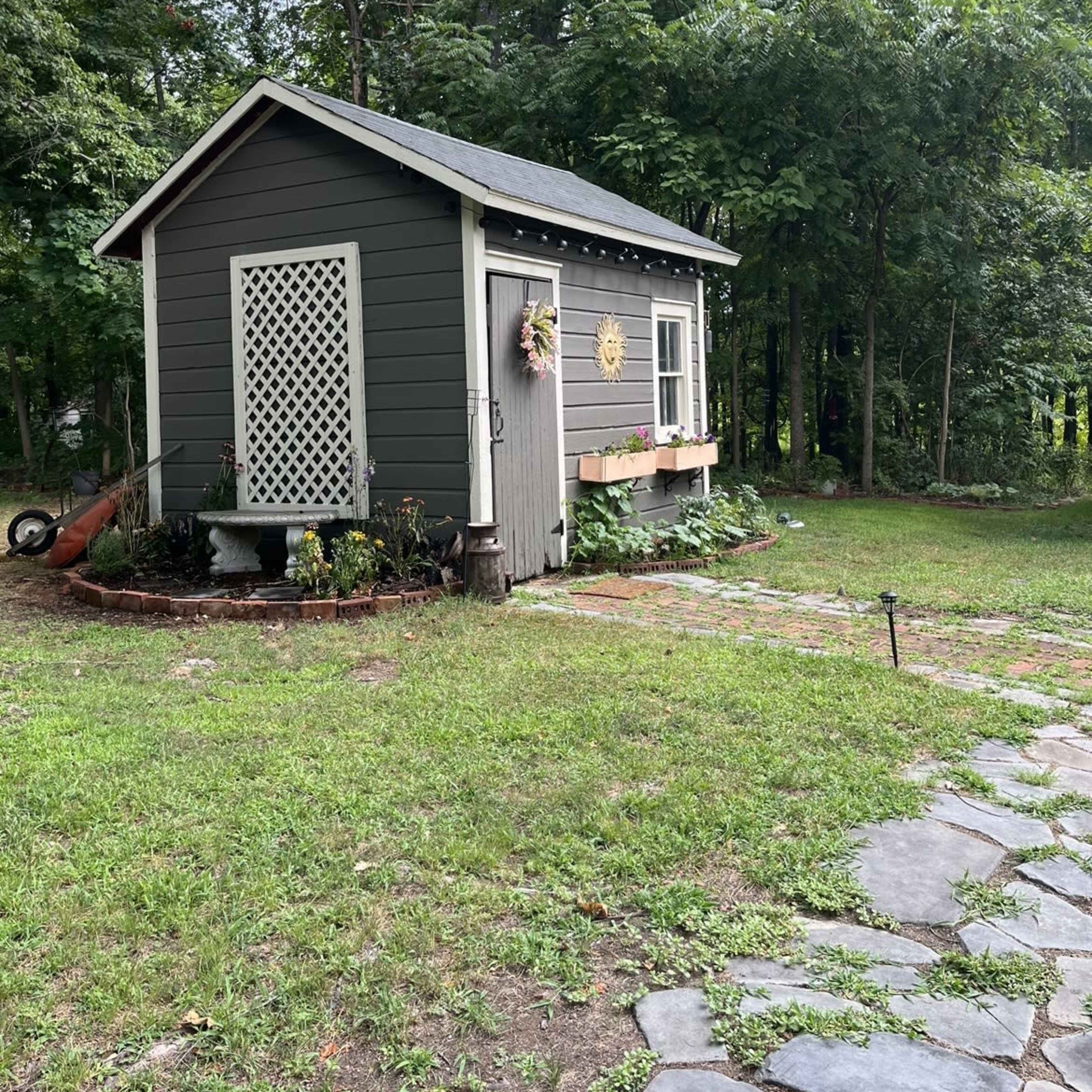 A small gray wooden shed with a lattice door is surrounded by a stone pathway and greenery.