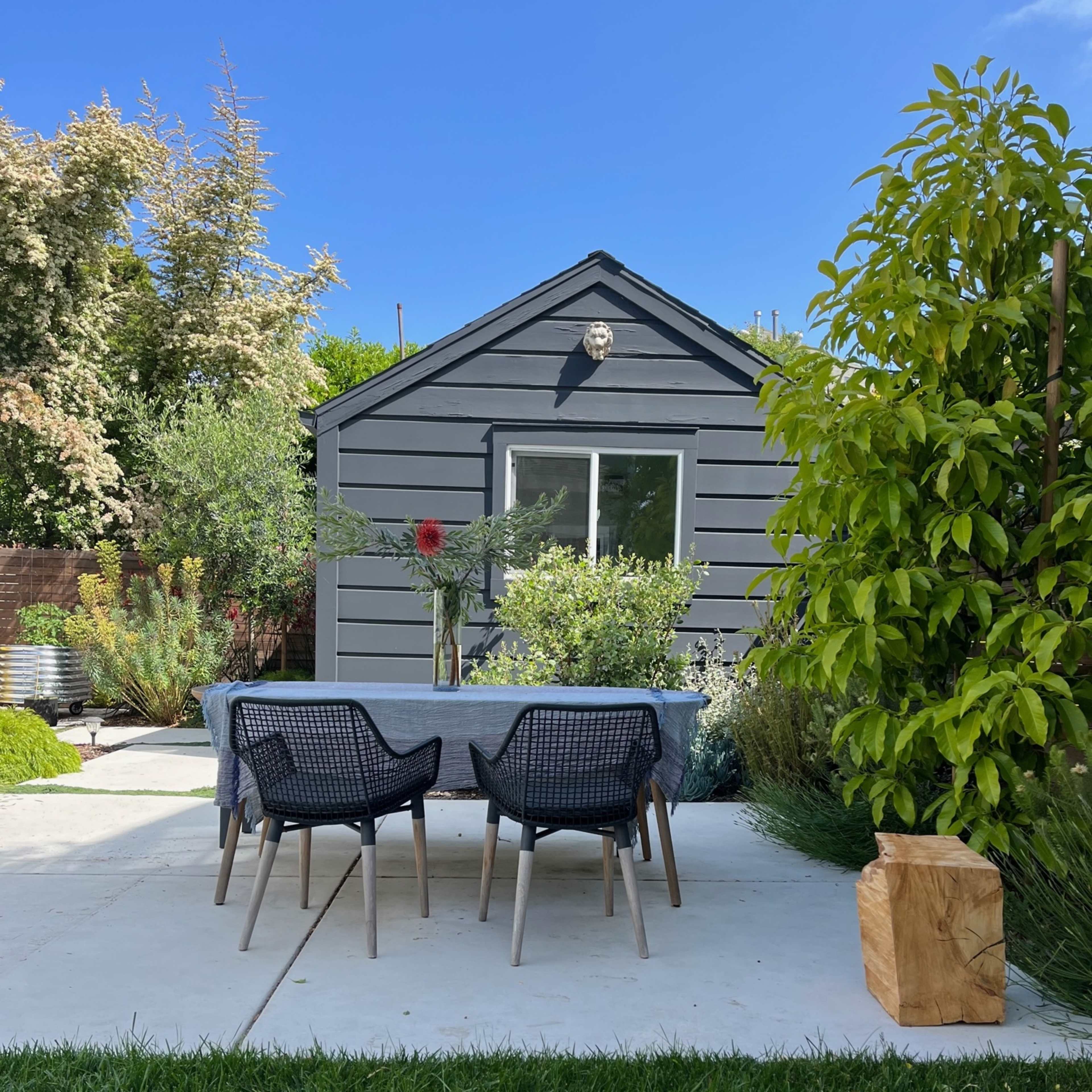 A gray cottage with a landscaped garden is visible in the background, featuring a table and four chairs set on a concrete patio.