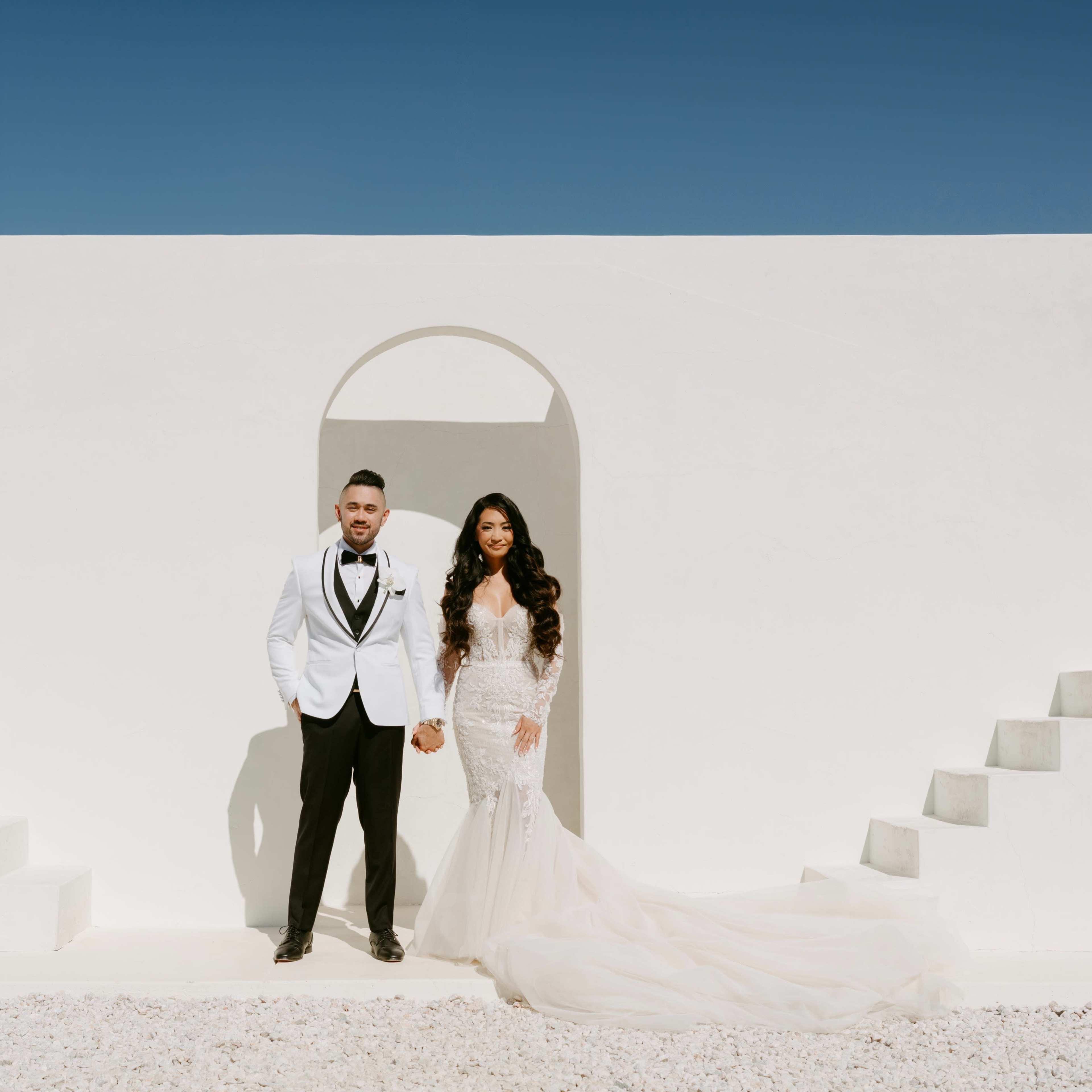 A bride and groom stand hand in hand in front of a minimalist white structure with stairs leading up, under a clear blue sky.