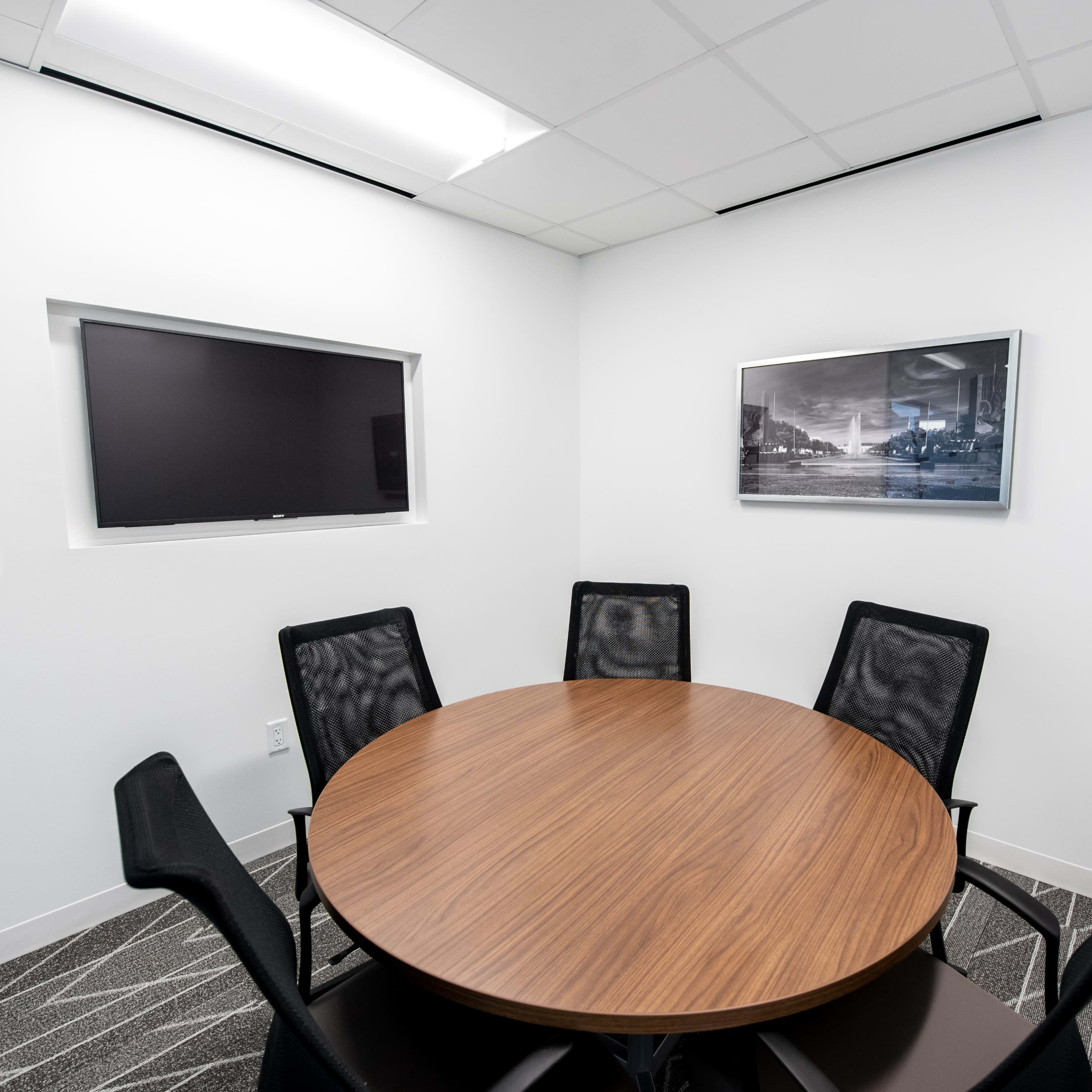 A modern conference room with a round wooden table surrounded by five black chairs, featuring a wall-mounted television and a framed photograph.