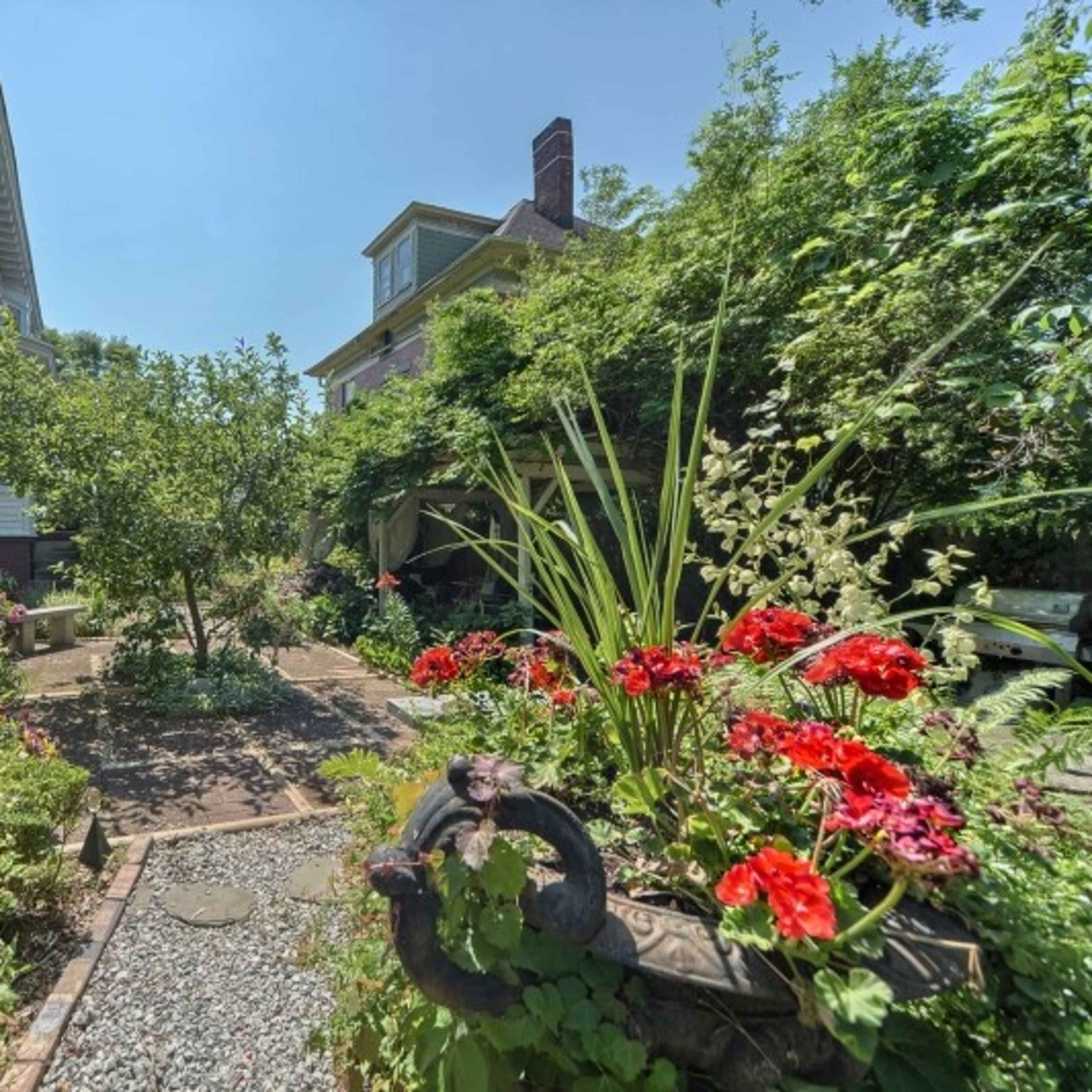 A garden pathway is flanked by vibrant flower beds and greenery, leading to two large houses in the background.