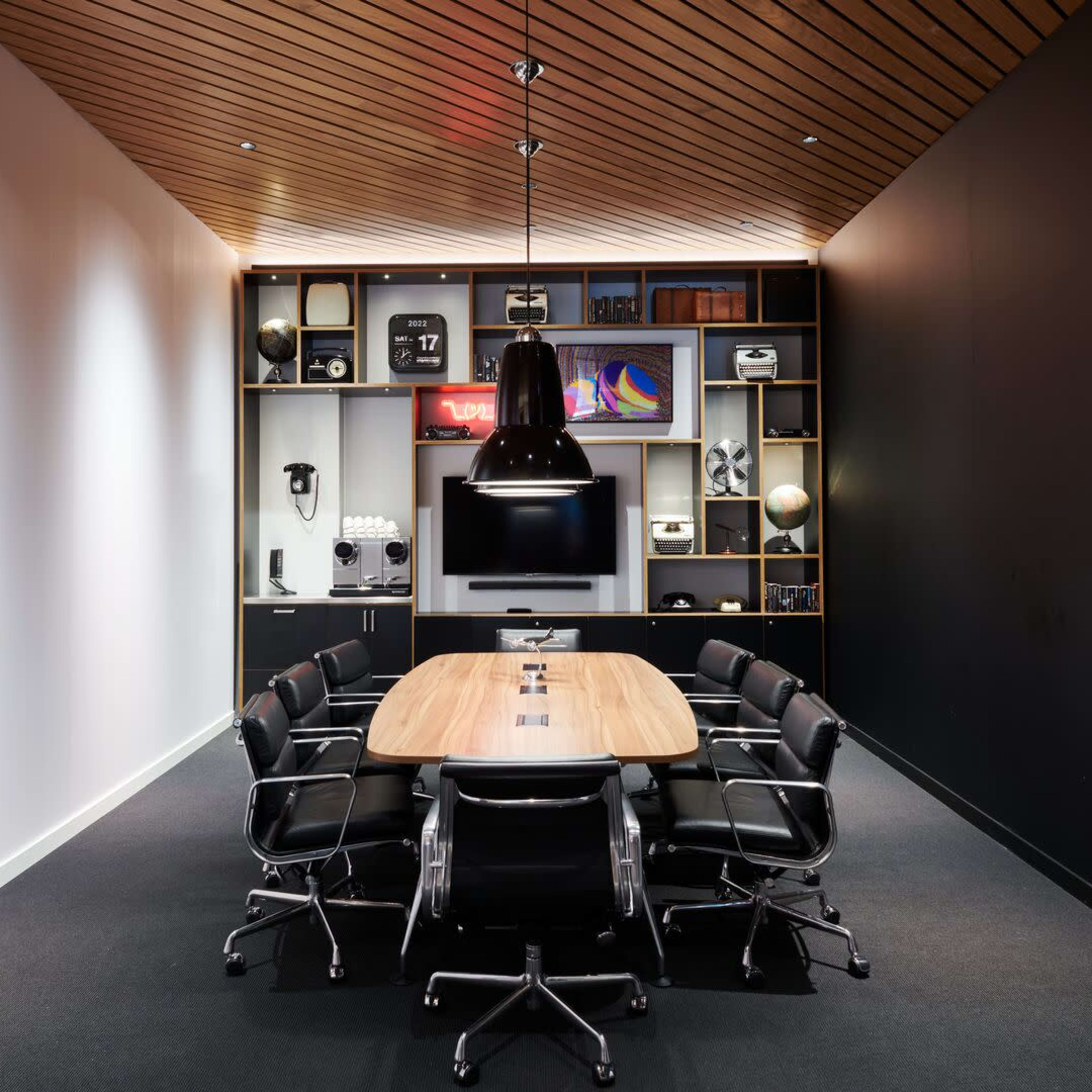 A modern conference room features a long wooden table surrounded by black chairs, with a multimedia setup and a wall of shelves displaying various items.