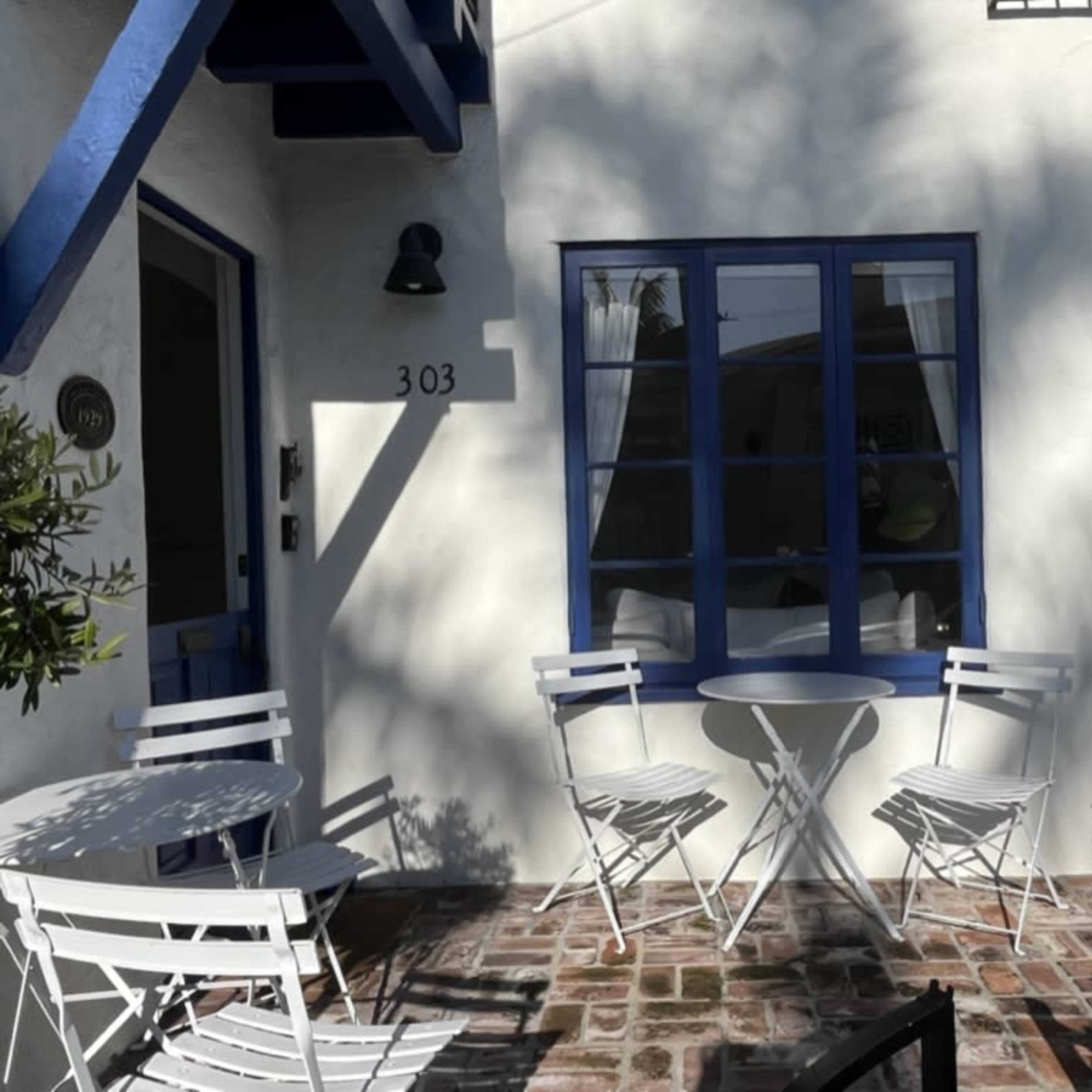 A small outdoor seating area with white metal chairs and a round table in front of a building featuring a blue window and door.
