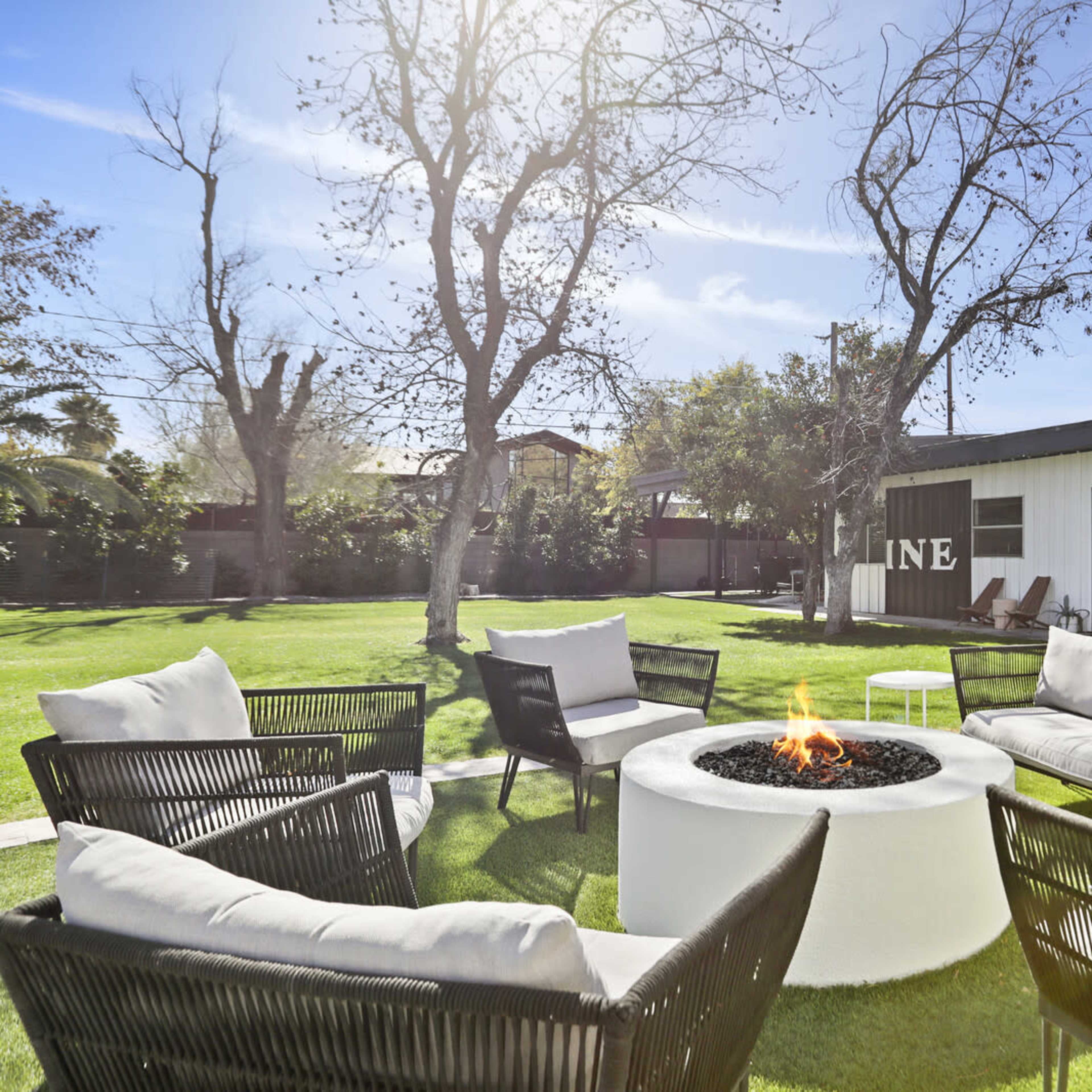 A circular fire pit surrounded by black chairs and white couches occupies a grassy backyard with trees and a modern building in the background.
