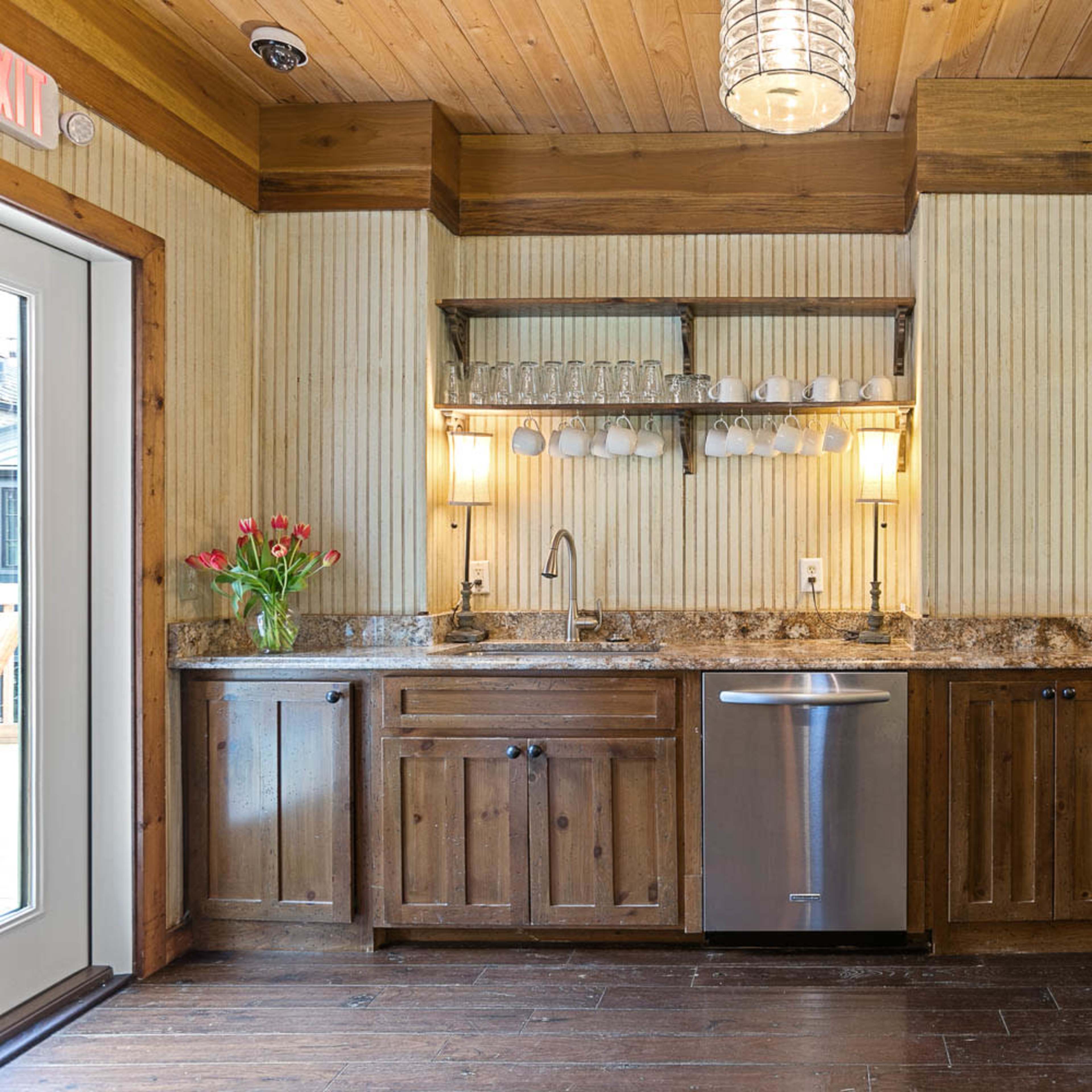 A wooden kitchen area with a countertop, cabinets, stainless steel dishwasher, and a shelf holding glasses.