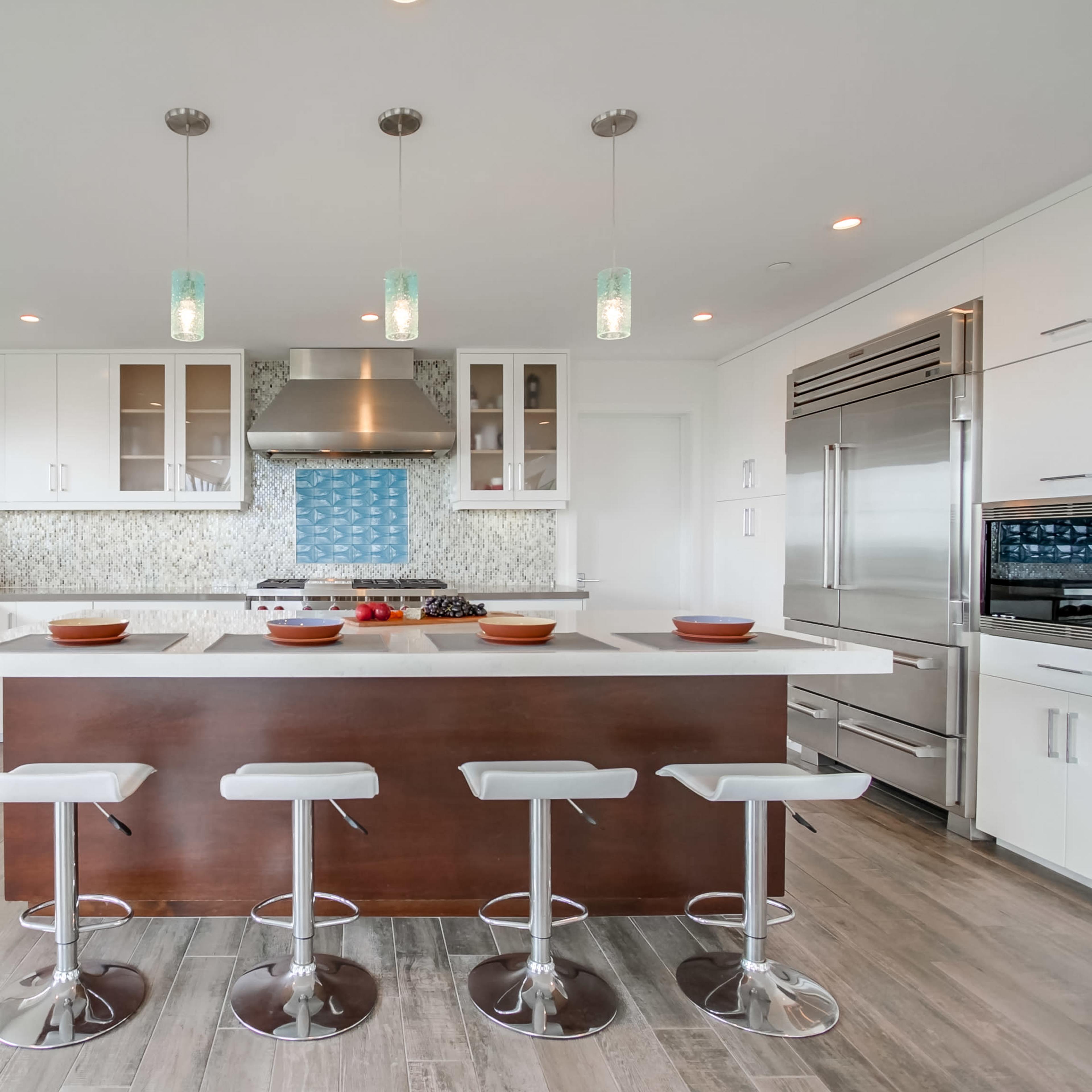 A modern kitchen featuring a large island with four white stools, stainless steel appliances, and light-colored cabinetry against a patterned backsplash.
