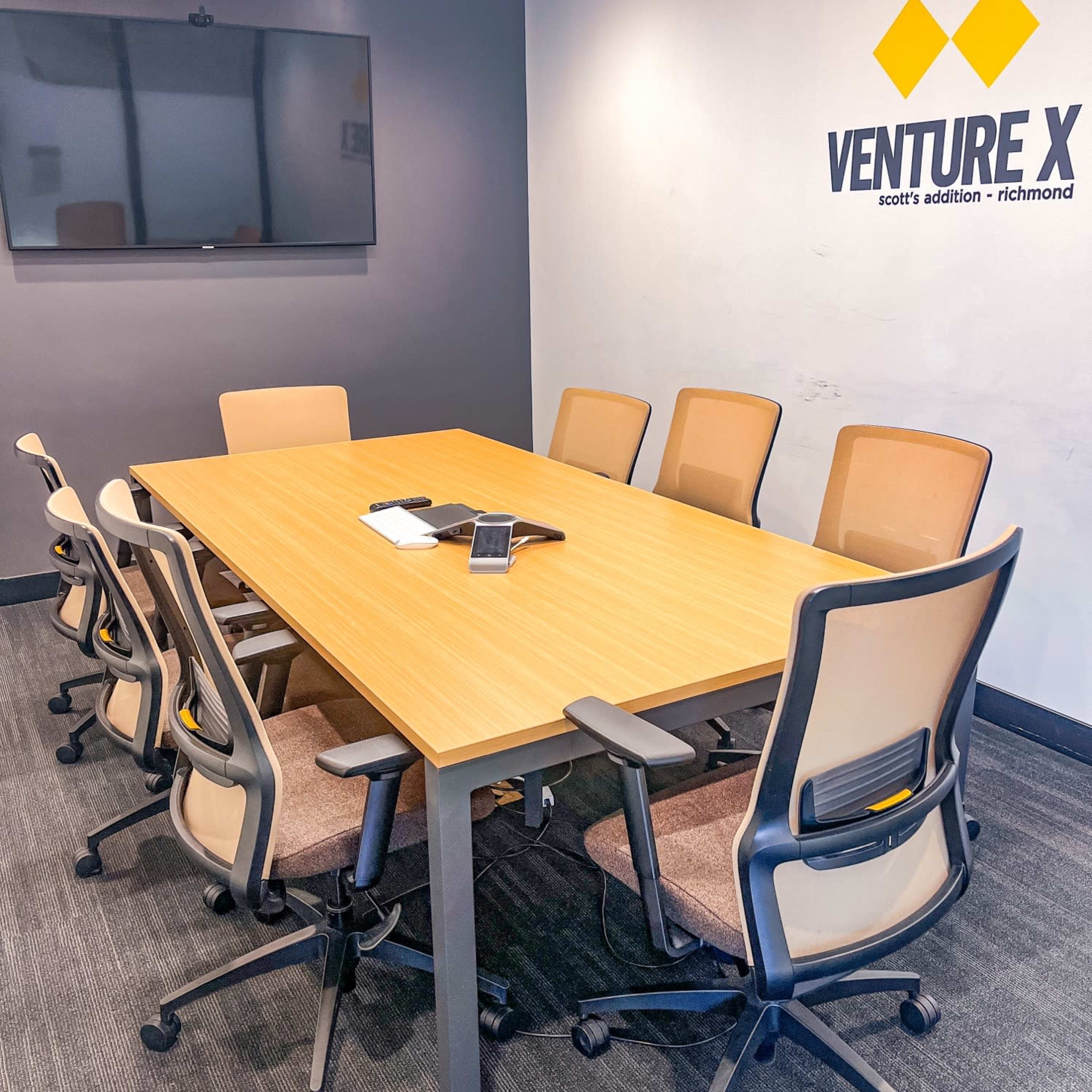 A modern conference room with a large wooden table surrounded by eight ergonomic chairs and a wall-mounted television.