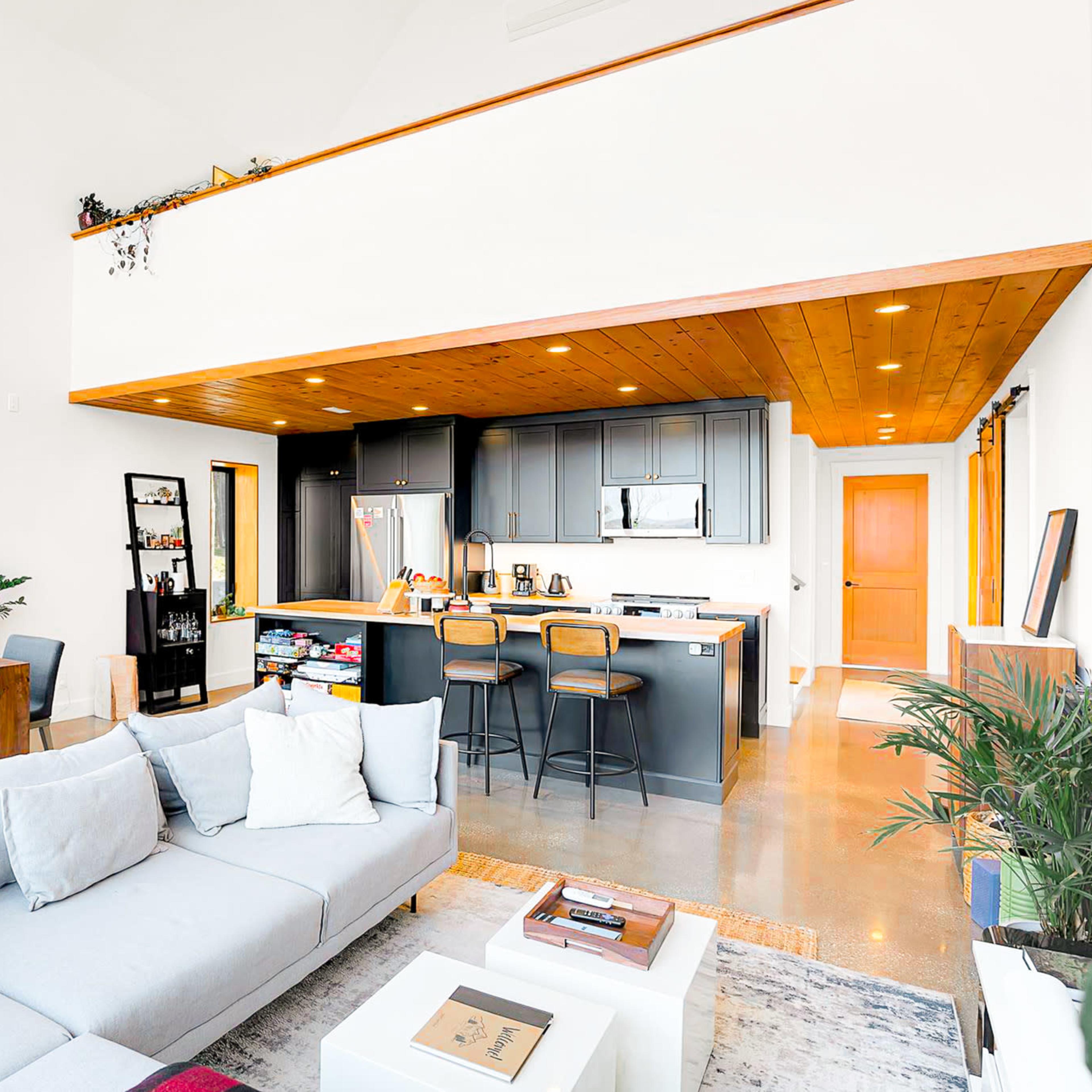 A modern living space featuring a light grey sectional sofa, a dining area with a table and chairs, and a kitchen with dark grey cabinetry and stainless steel appliances, all illuminated by natural light from large windows.