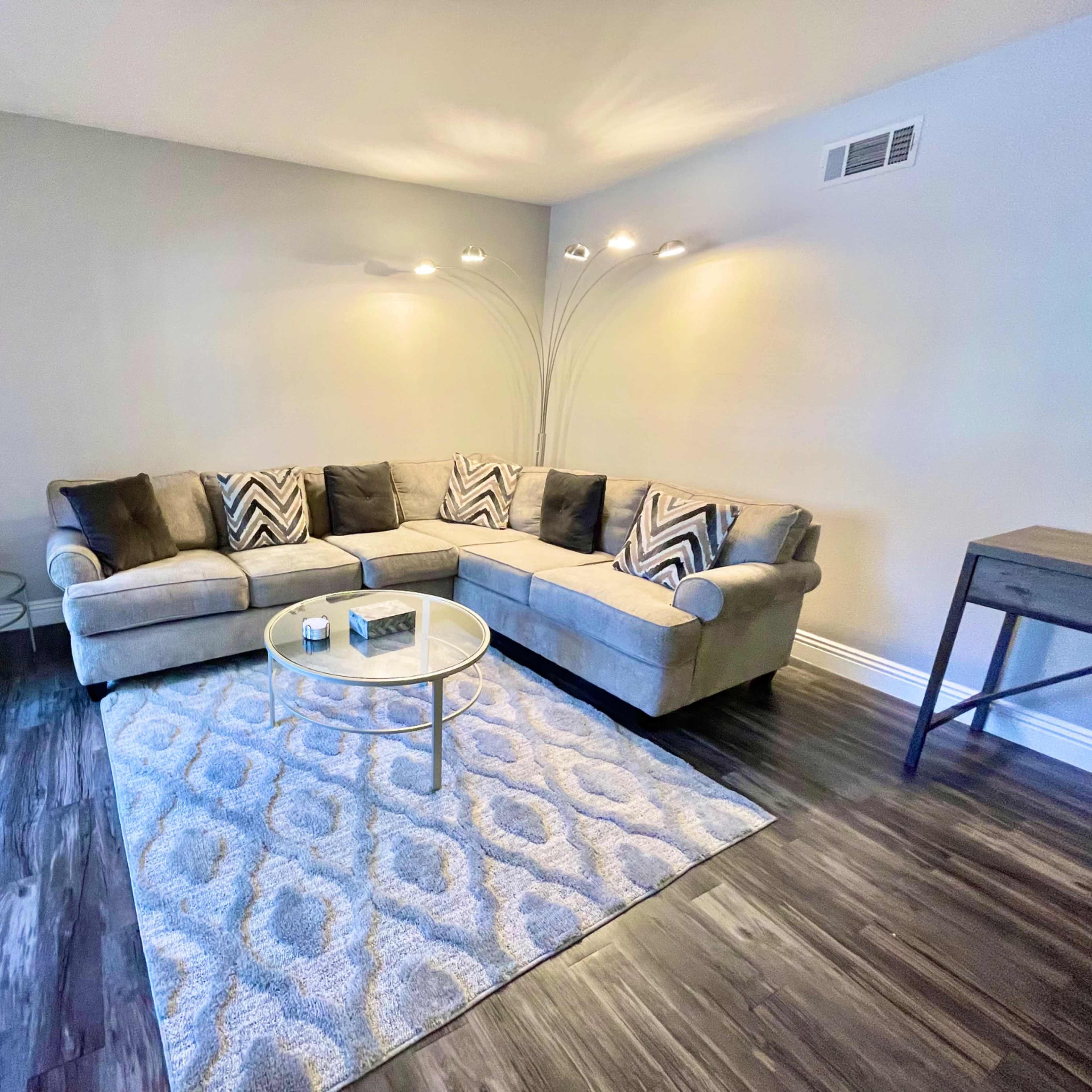 A living room with a sectional sofa, decorative throw pillows, a round glass coffee table, and a patterned rug on a wooden floor.