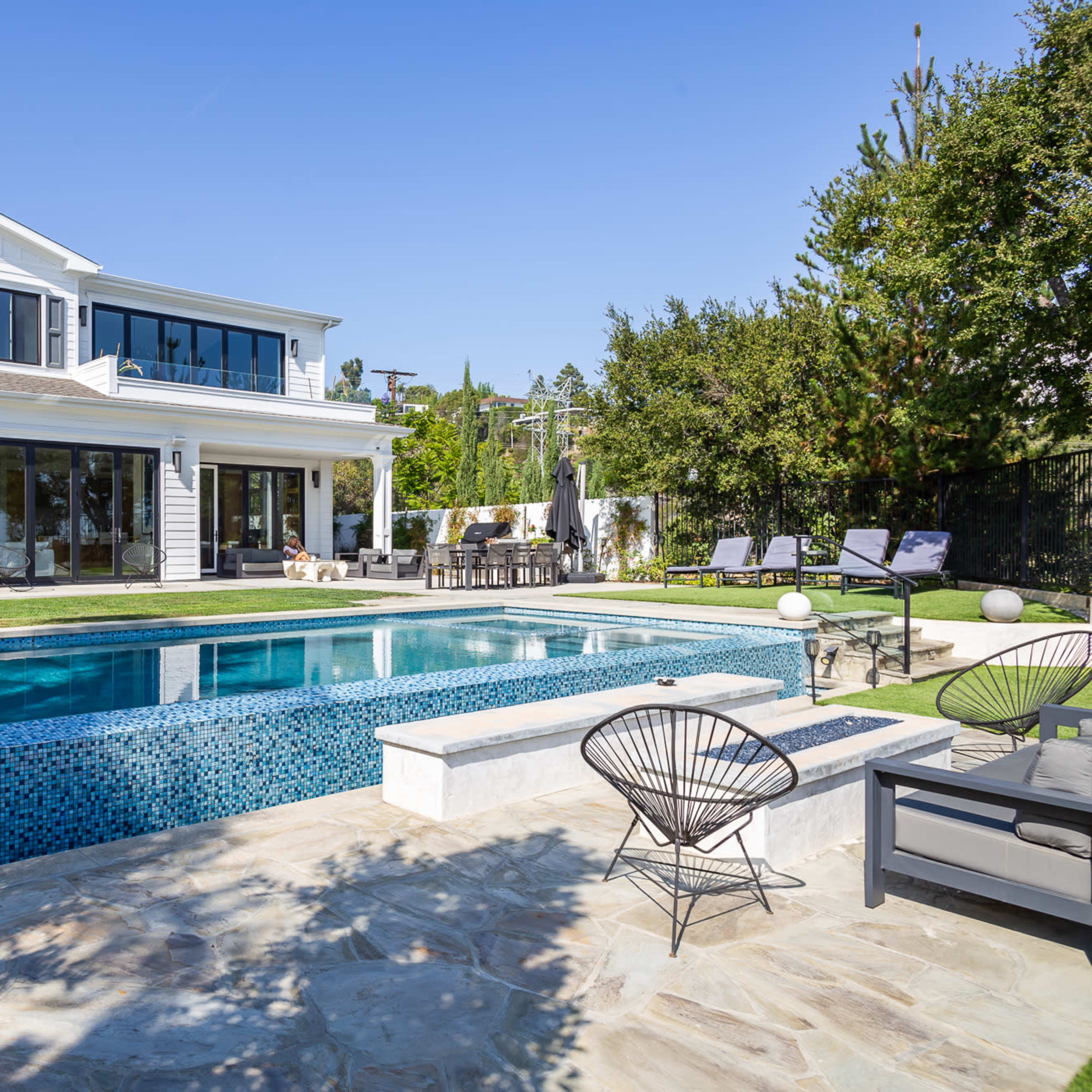 A modern house with large windows overlooks a swimming pool surrounded by lounge chairs and a patio area.