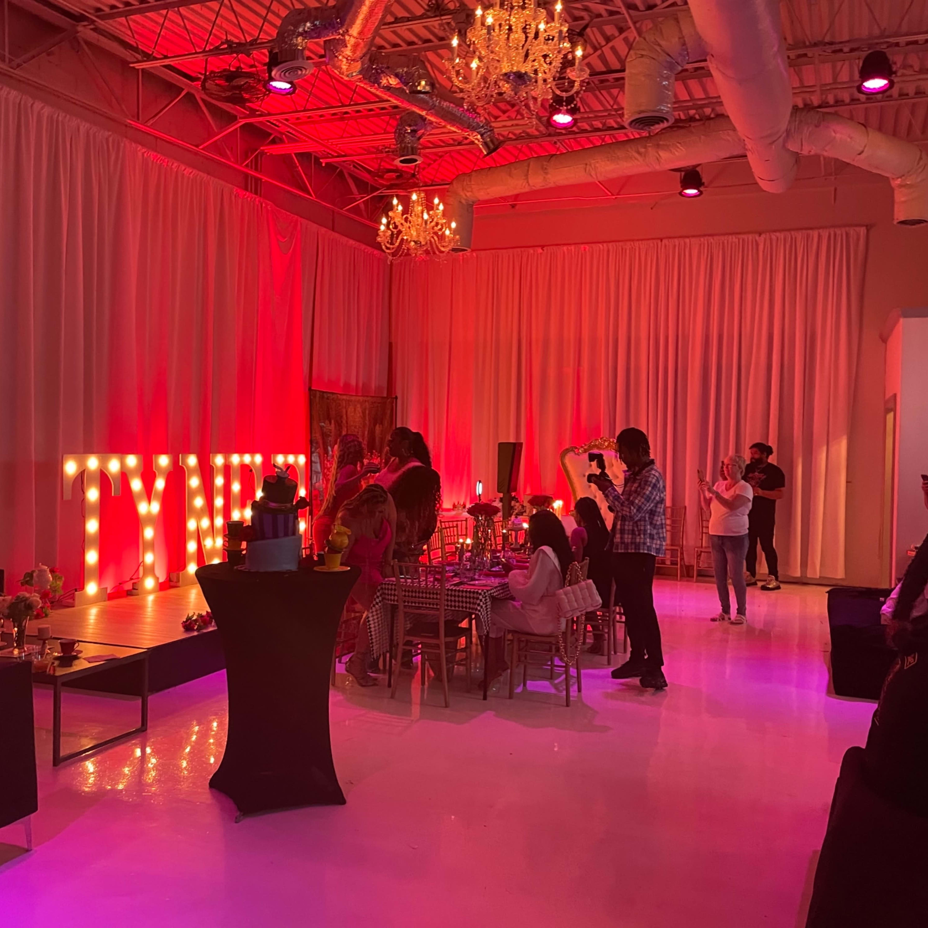 The scene shows a lively event space decorated with large illuminated letters spelling "TYME," featuring tables, guests mingling, and chandeliers hanging from the ceiling.