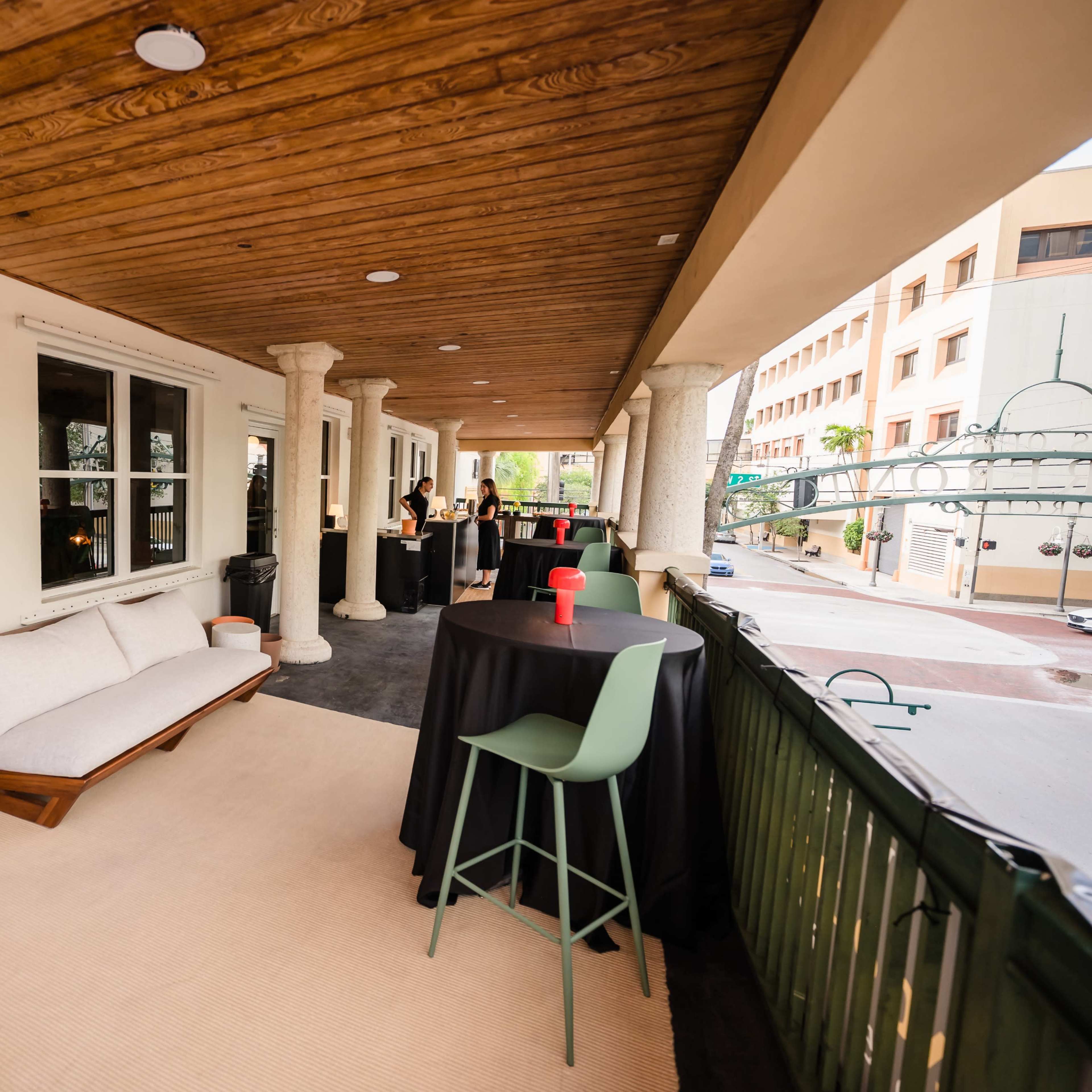 A covered outdoor patio area featuring a couch, tall tables with chairs, and a view of a building exterior.