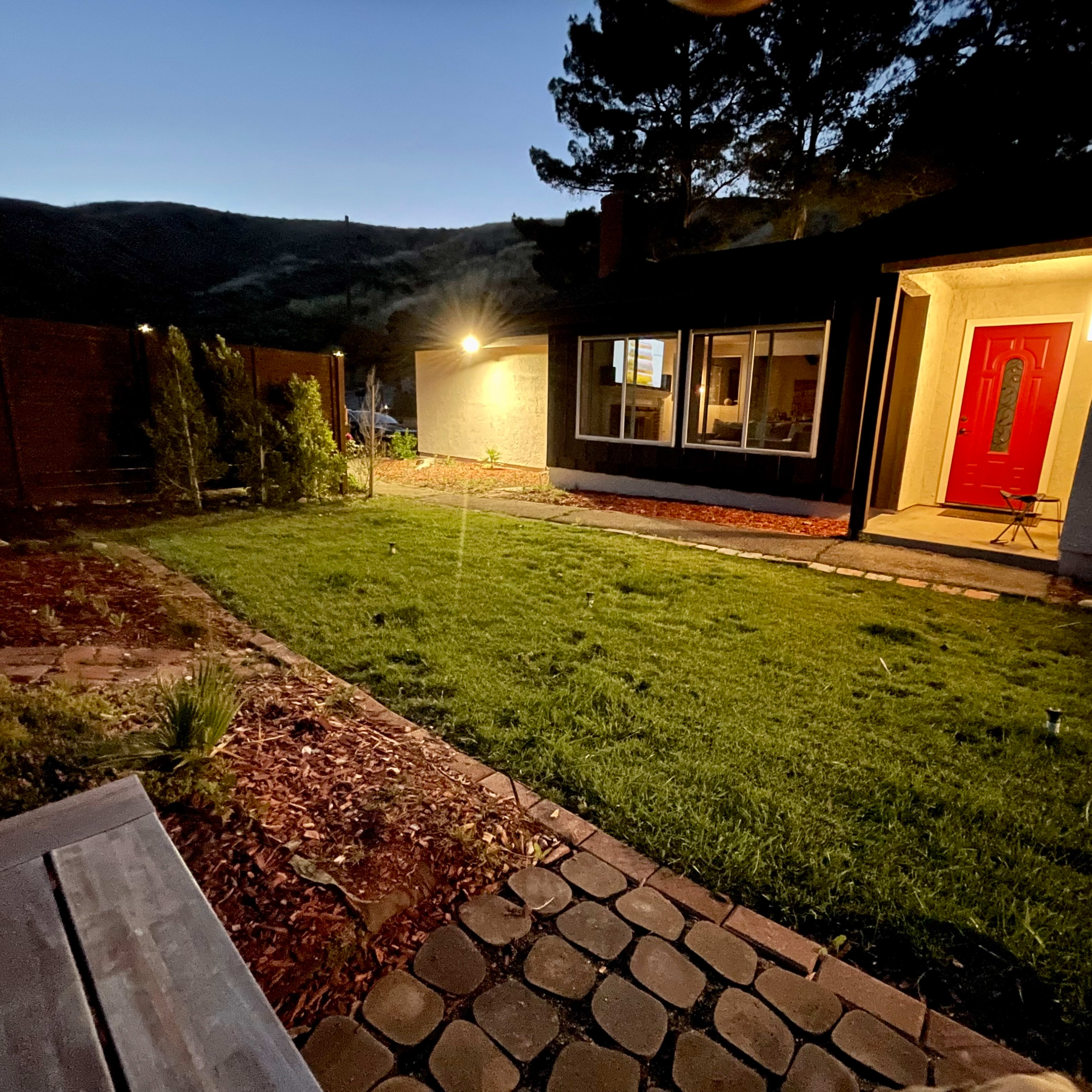 A well-maintained backyard features a lawn and stone pathway, illuminated by a porch light under a twilight sky.
