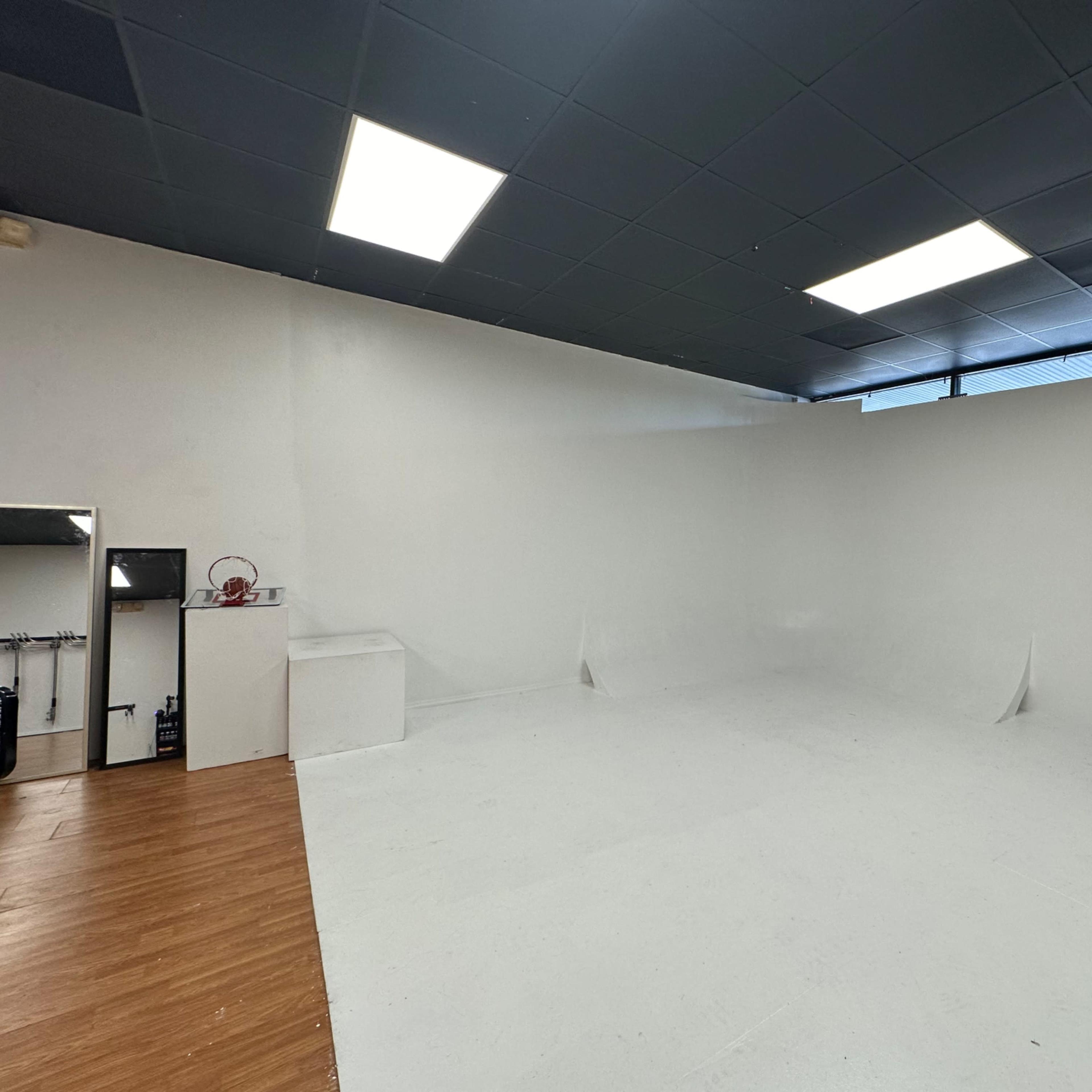 A spacious, empty studio with white walls and a light-colored floor, featuring a small area with equipment and a black curtain in one corner.