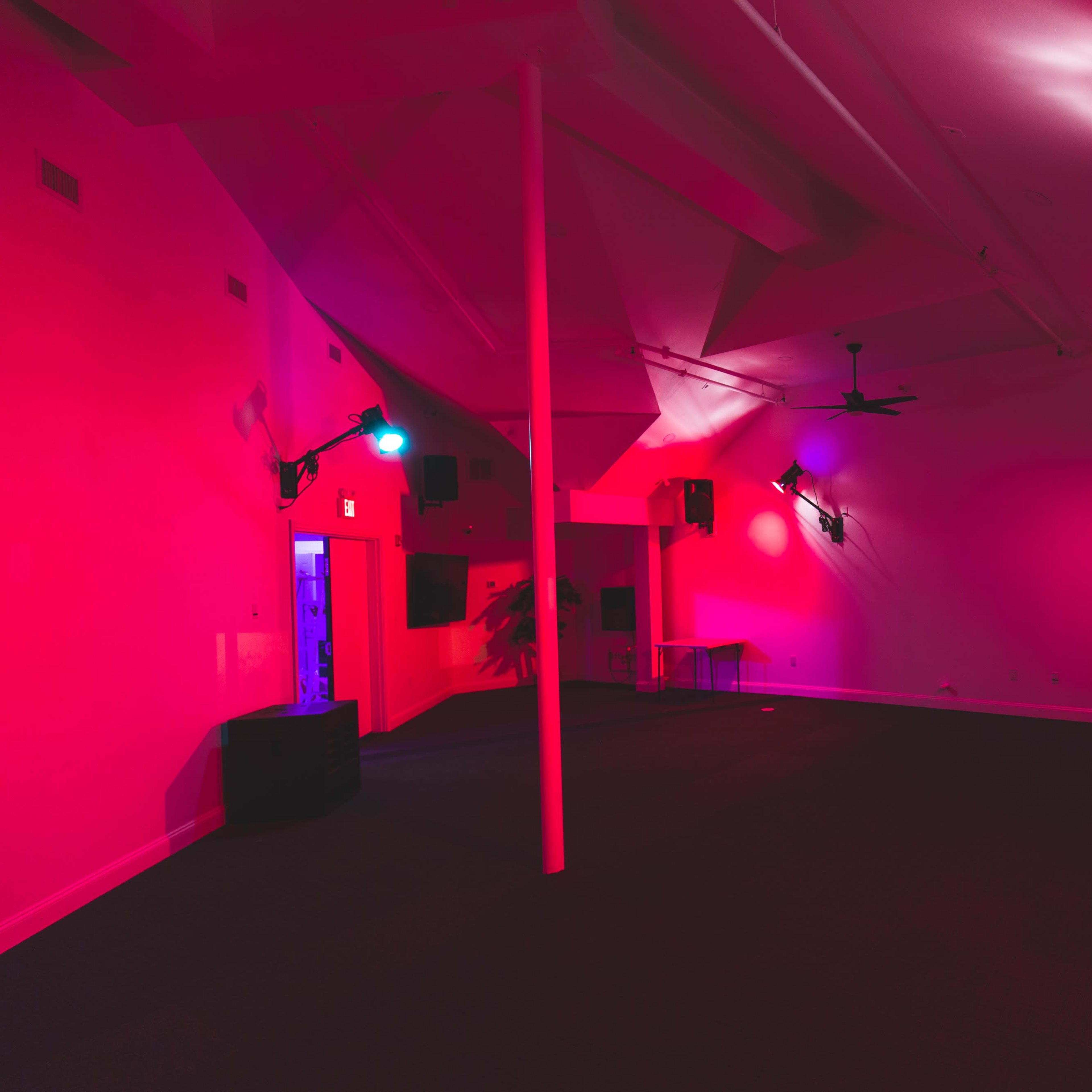A dimly lit room with pink and blue lighting, featuring several wall-mounted lights and a few bar stools along one side.
