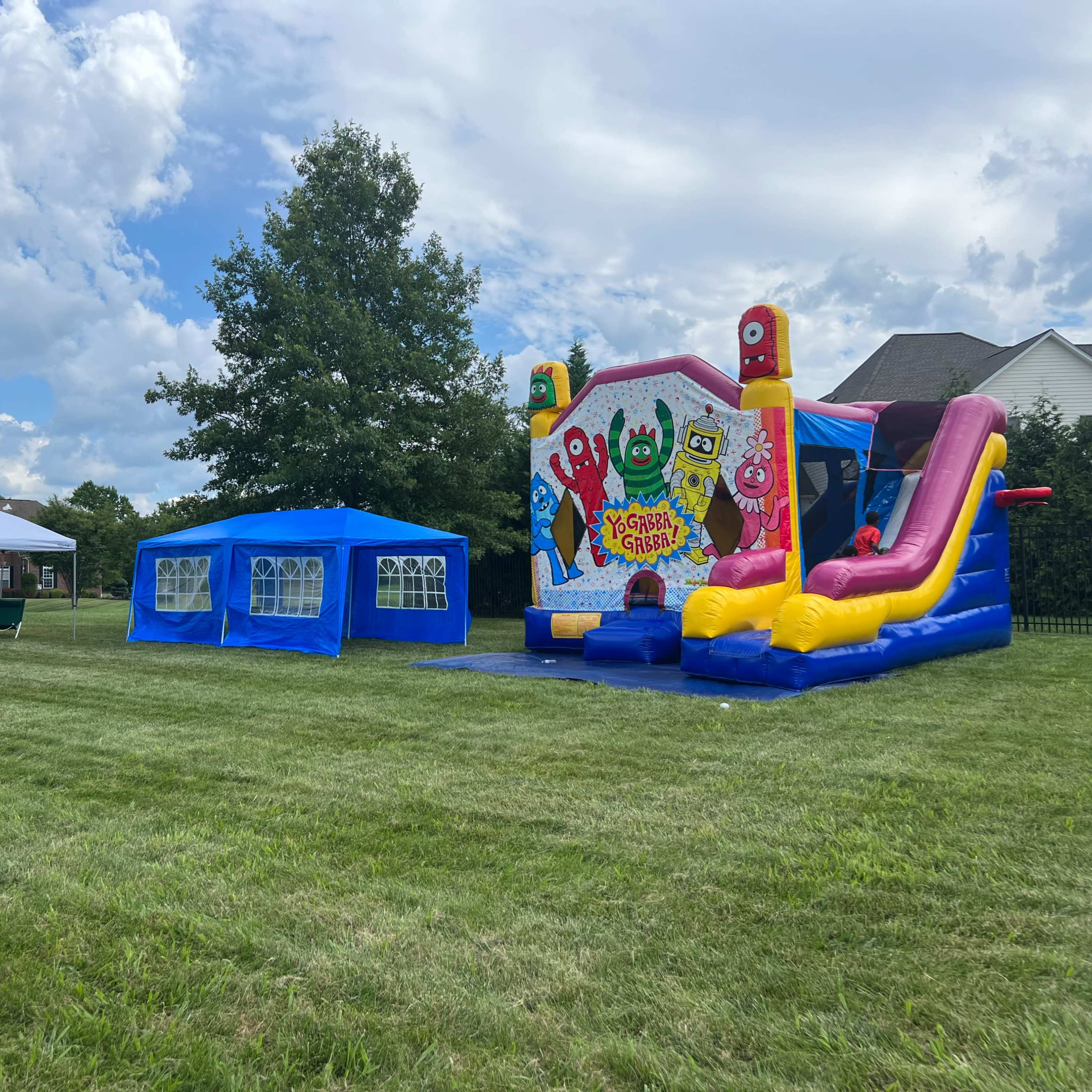 A colorful bounce house with slides is set up beside a blue tent and seating area on a green lawn near a residential home.