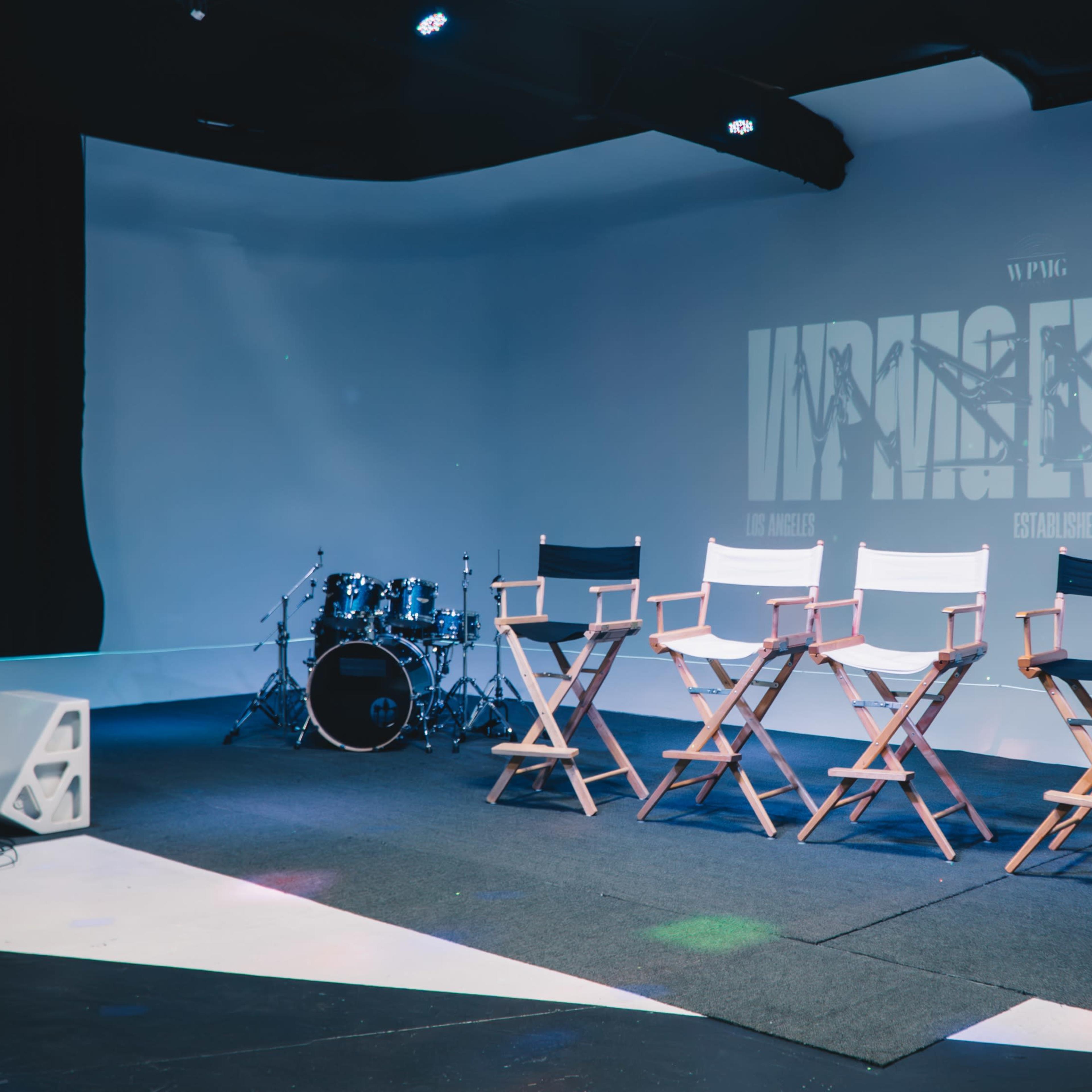A minimalist stage setup with three director's chairs, a drum set, and audio equipment against a white backdrop featuring the words "WINGS EVENTS."
