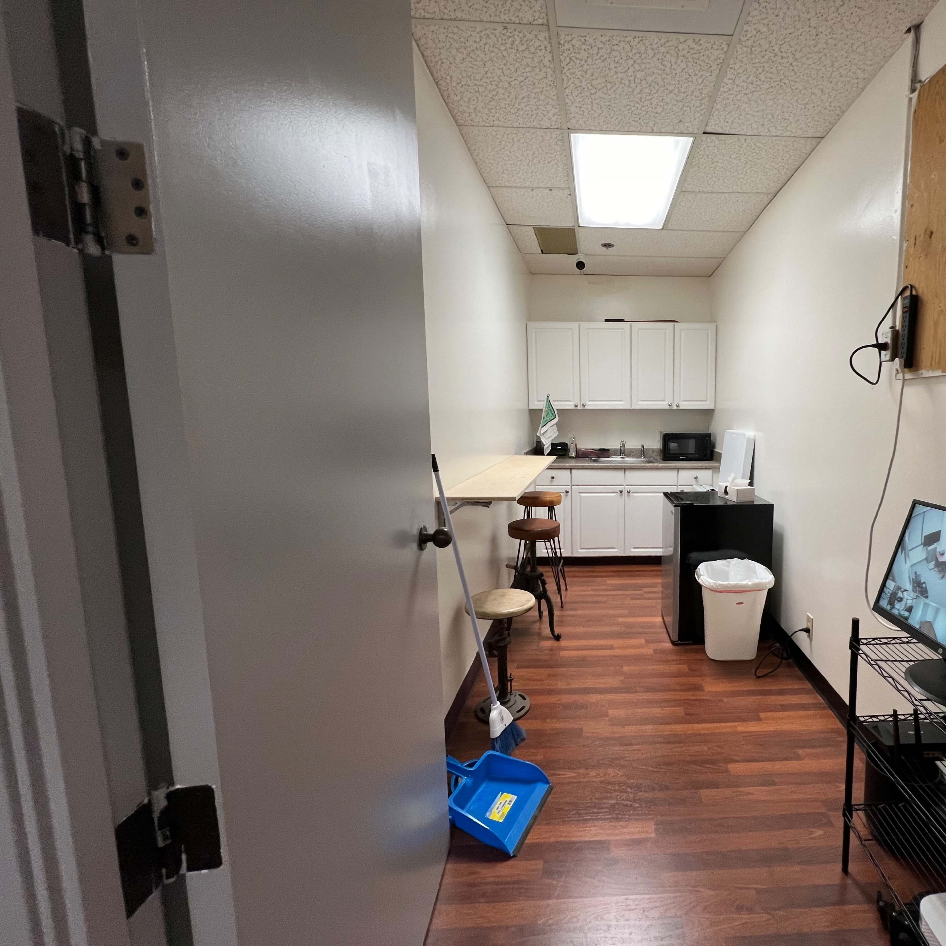 A narrow room with a kitchenette on one side, a small table with two stools, a trash can, and a TV displaying security footage.