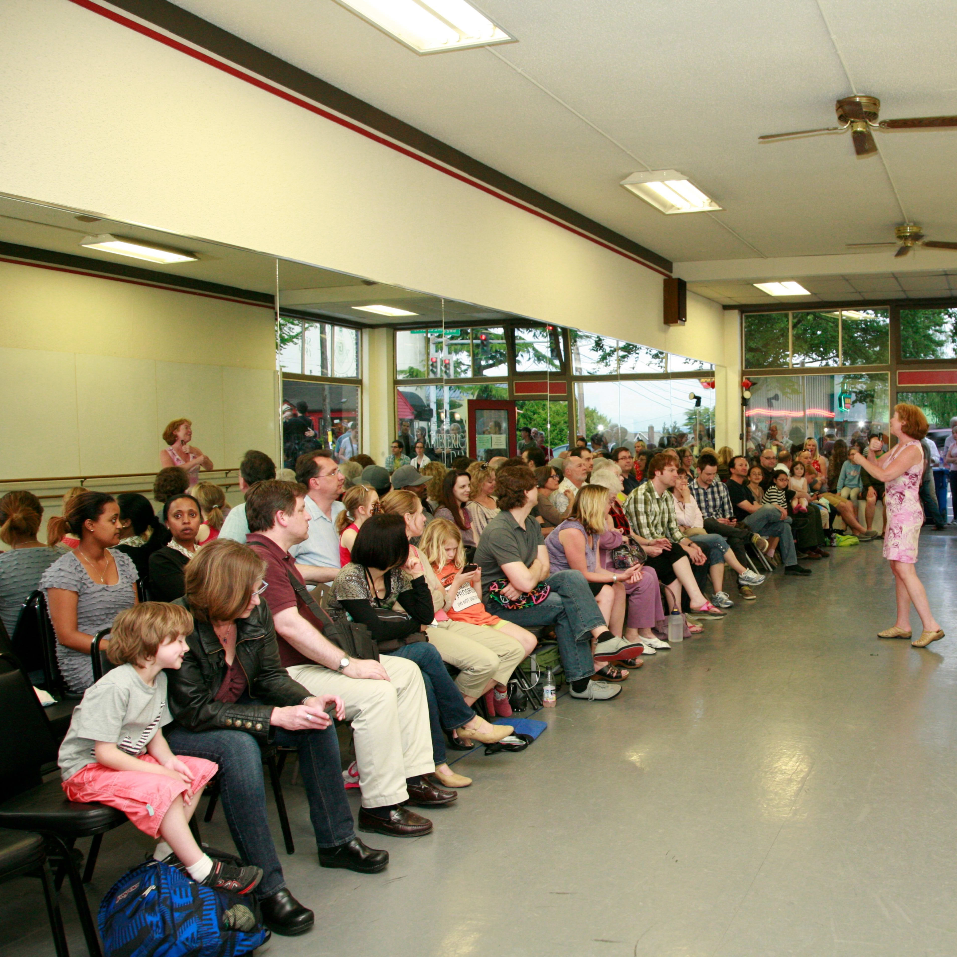 A crowd of people sits in a dance studio with mirrors and a wooden floor, while a person stands and speaks at the front.