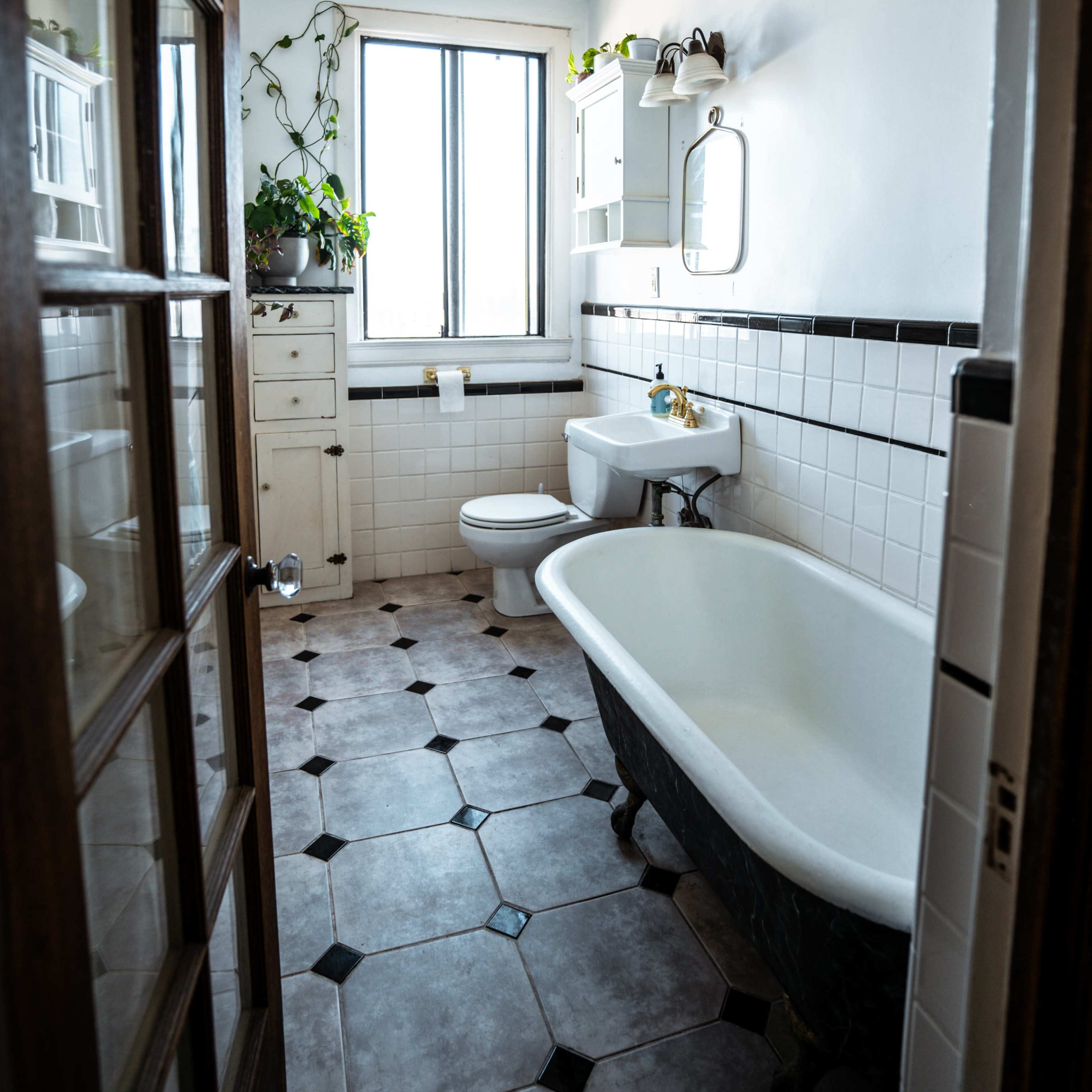 A bright bathroom features a freestanding bathtub, a toilet, a sink with a mirror, and several plants near the windows.