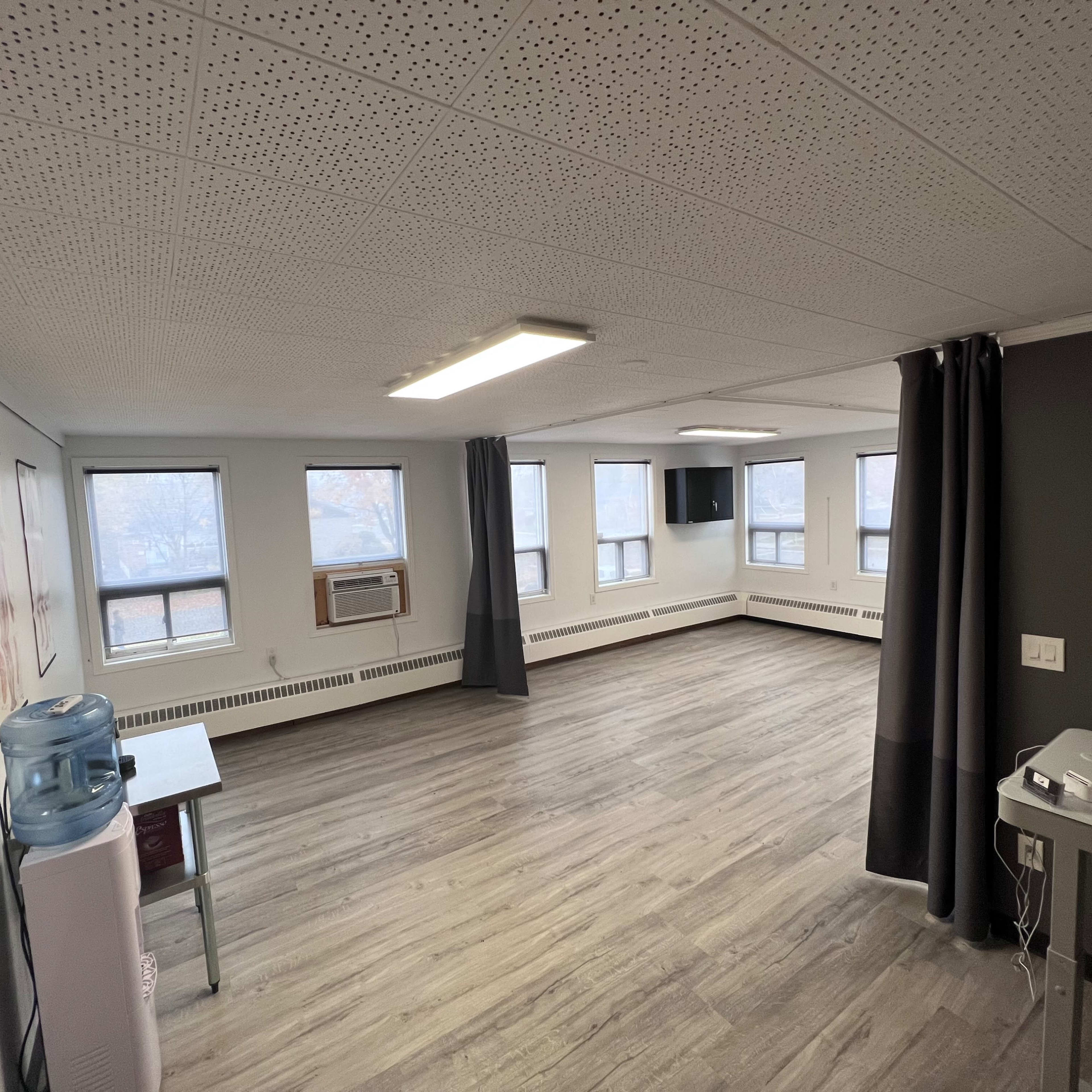 An empty room with wooden flooring, multiple windows, a wall-mounted television, and medical posters hanging on the walls.
