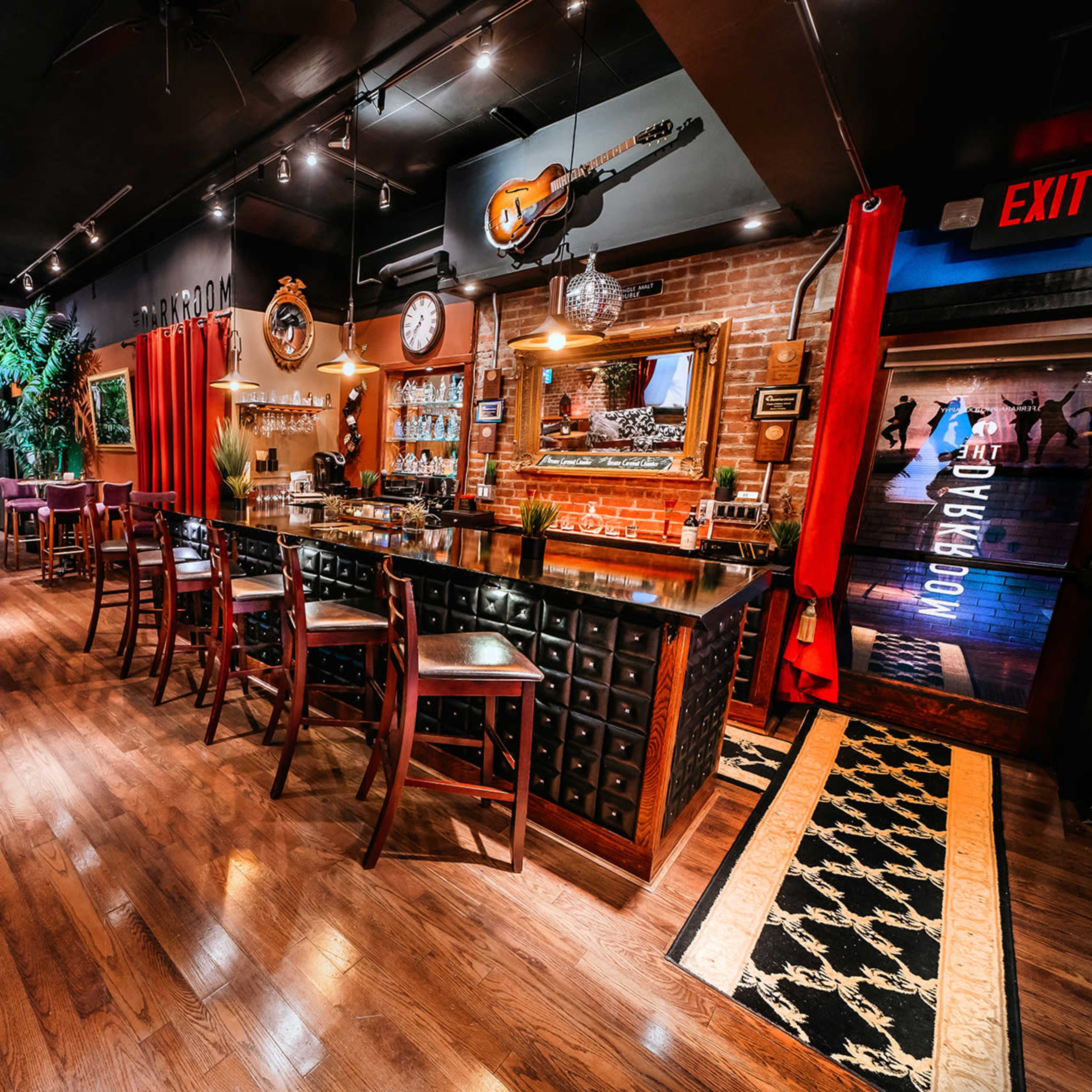 A dimly lit bar with wooden flooring, a counter lined with stools, and tables arranged throughout a cozy lounge area, decorated with musical instruments and plants.