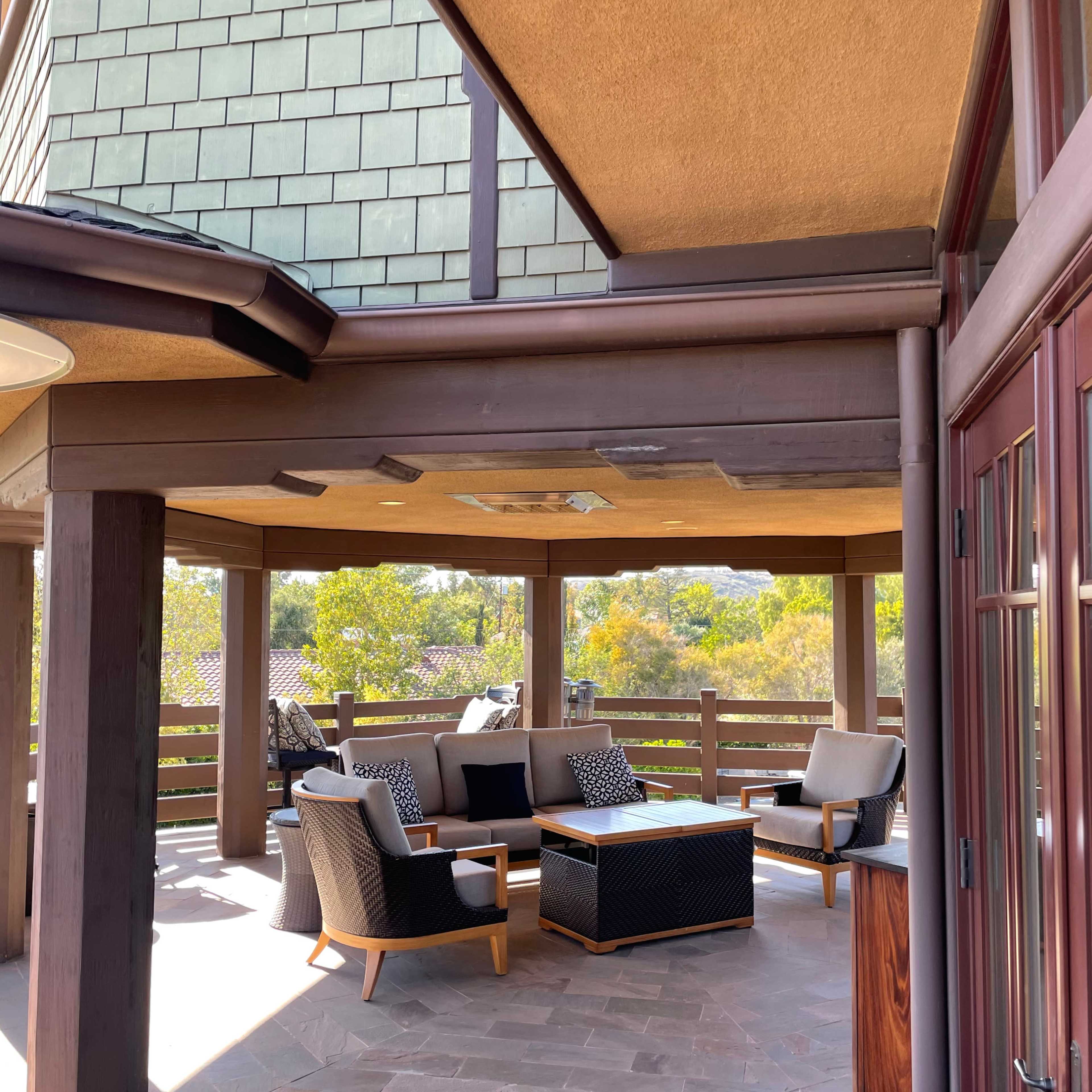 A covered outdoor patio area with seating and a coffee table, surrounded by wooden railings and greenery.