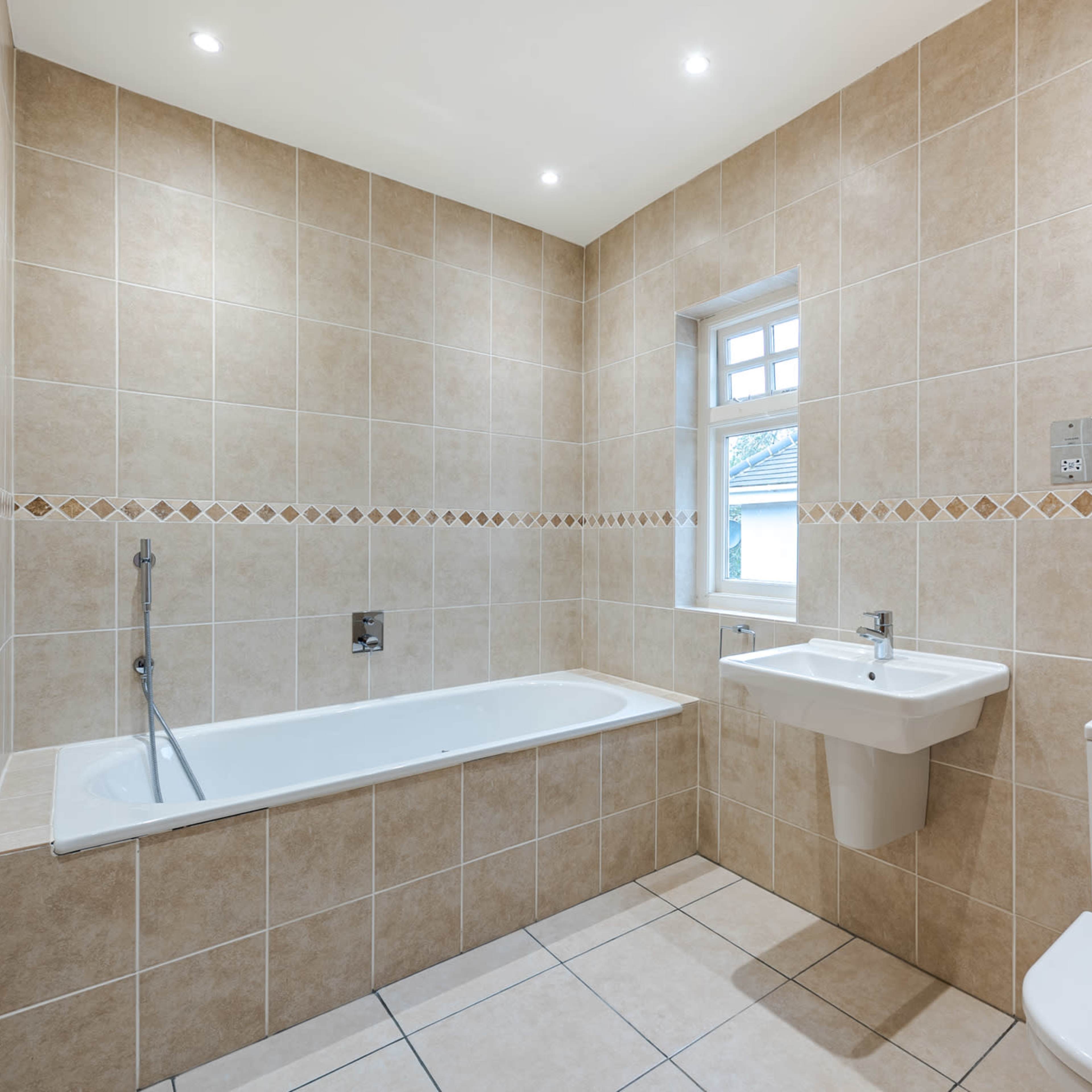 A bathroom featuring a bathtub, a sink, and a toilet, all tiled with beige tiles and illuminated by overhead lights.