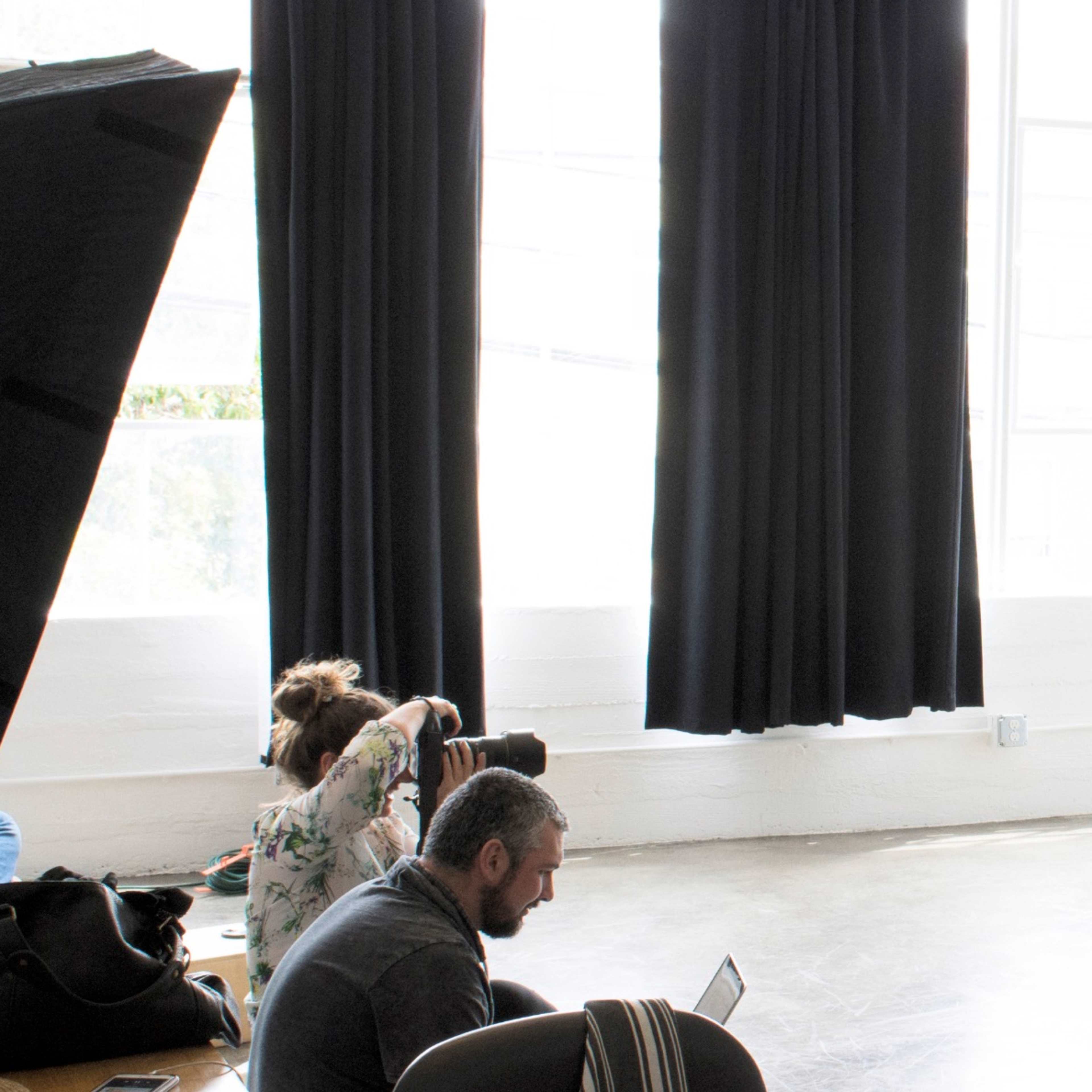 A child stands on a bare floor in a brightly lit studio space, while an audience sits on the left side, observing.
