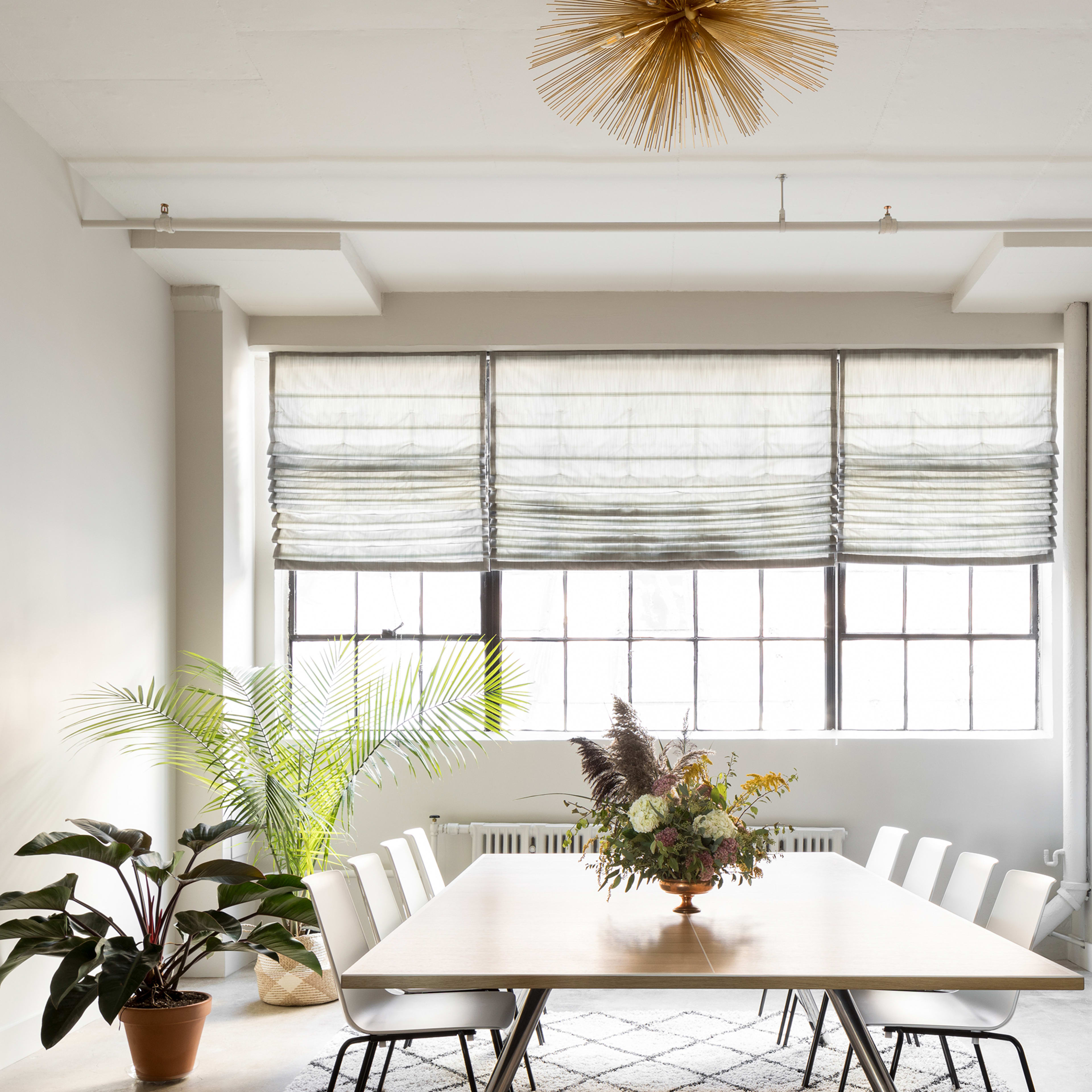 A modern dining room features a large table surrounded by white chairs, a decorative plant in the corner, and a television on a low cabinet beneath large windows with textured shades.