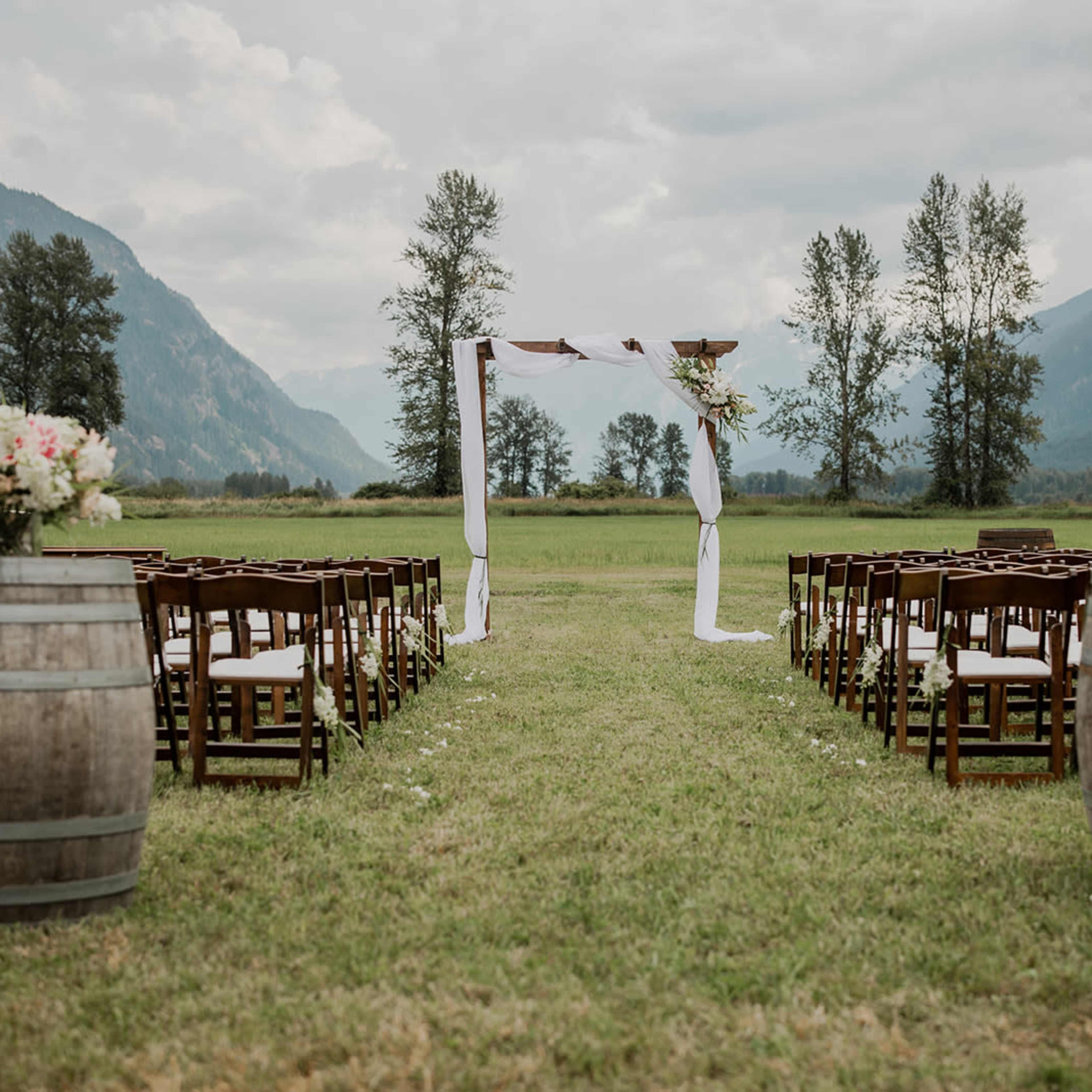 An outdoor wedding setting features rows of wooden chairs aligned toward a floral-decorated archway, with mountains and trees in the background.