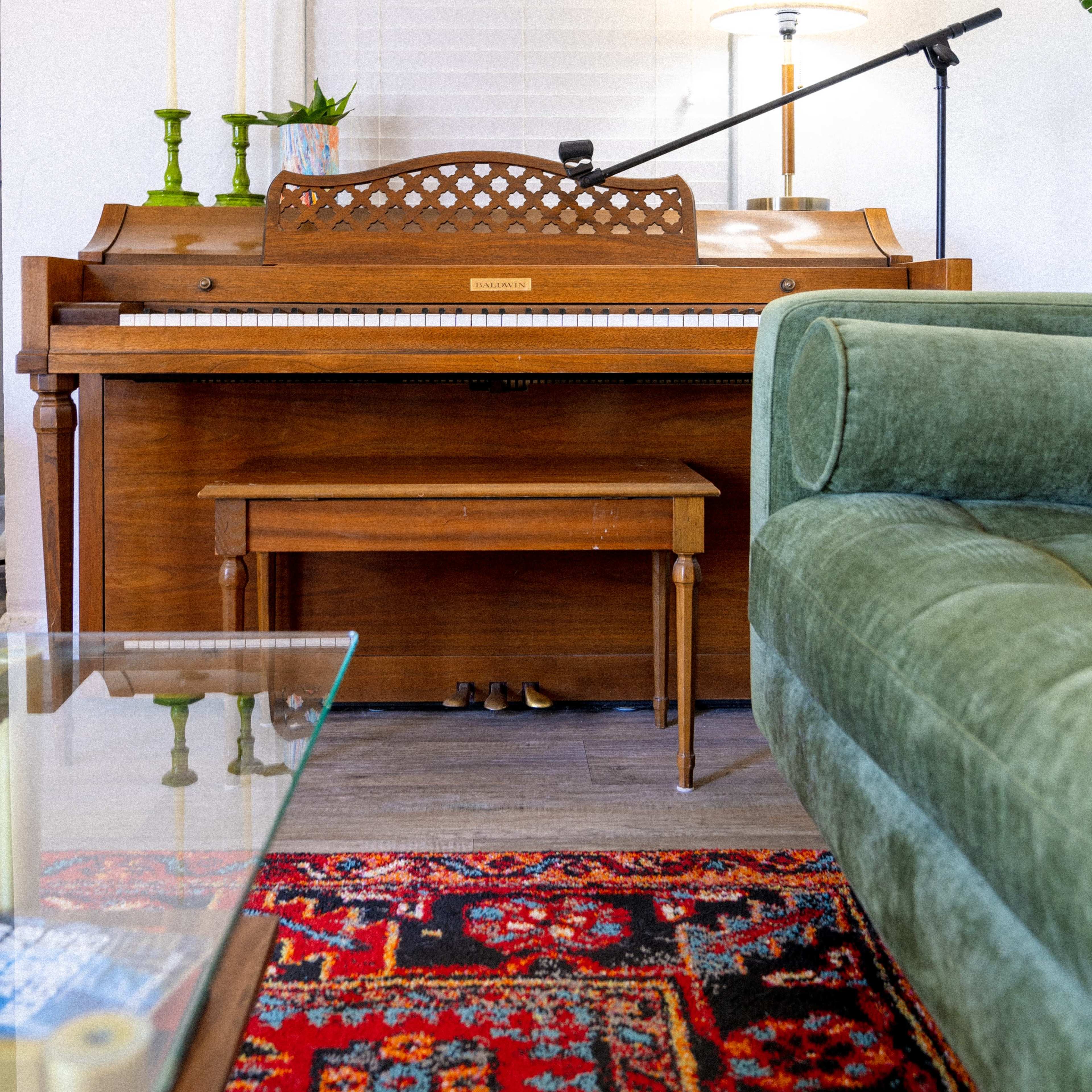 A wooden piano with a matching bench is positioned in a living room next to a green sofa and a lamp.