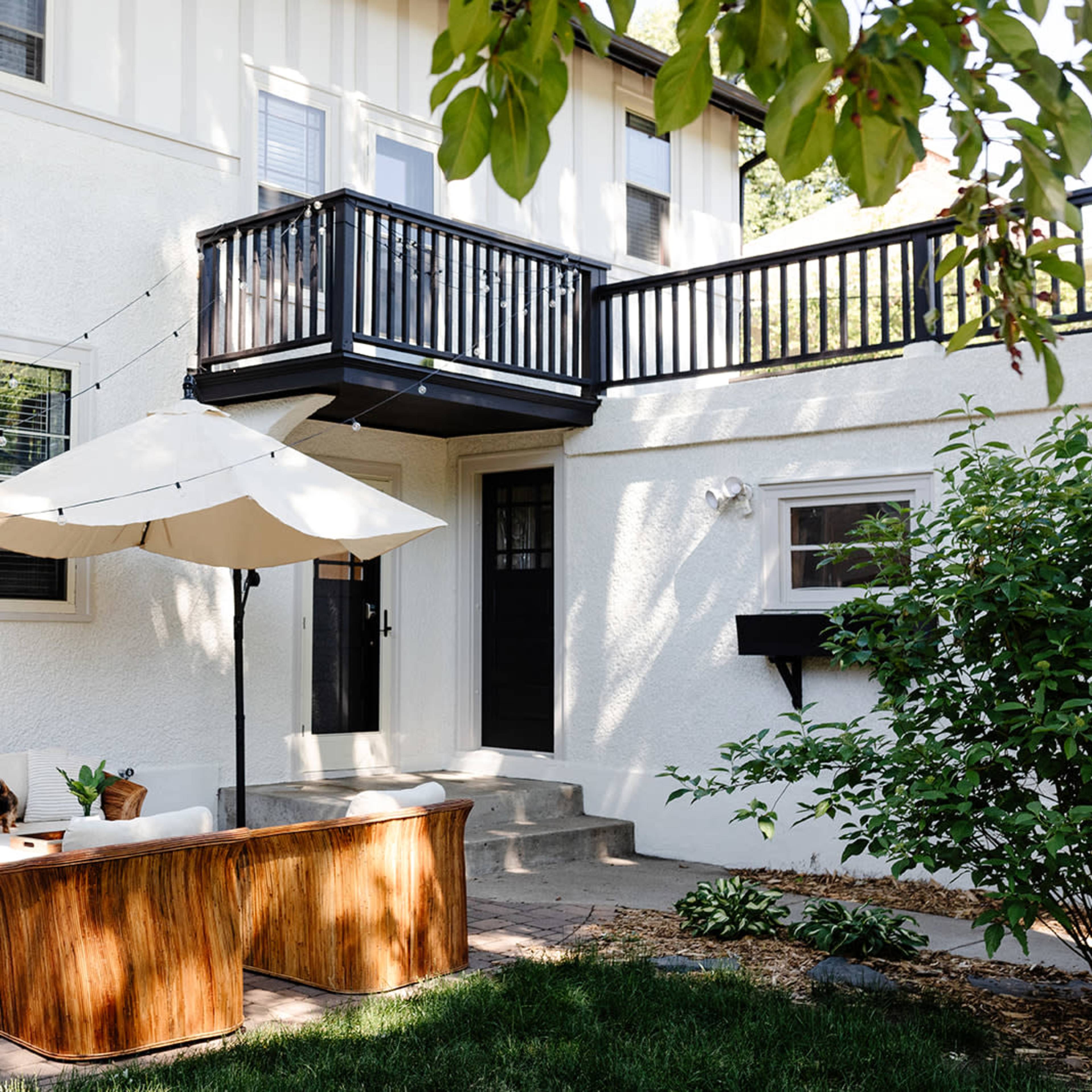 A patio area features a seating arrangement with a table, a large umbrella for shade, and an upper balcony overlooking a backyard.