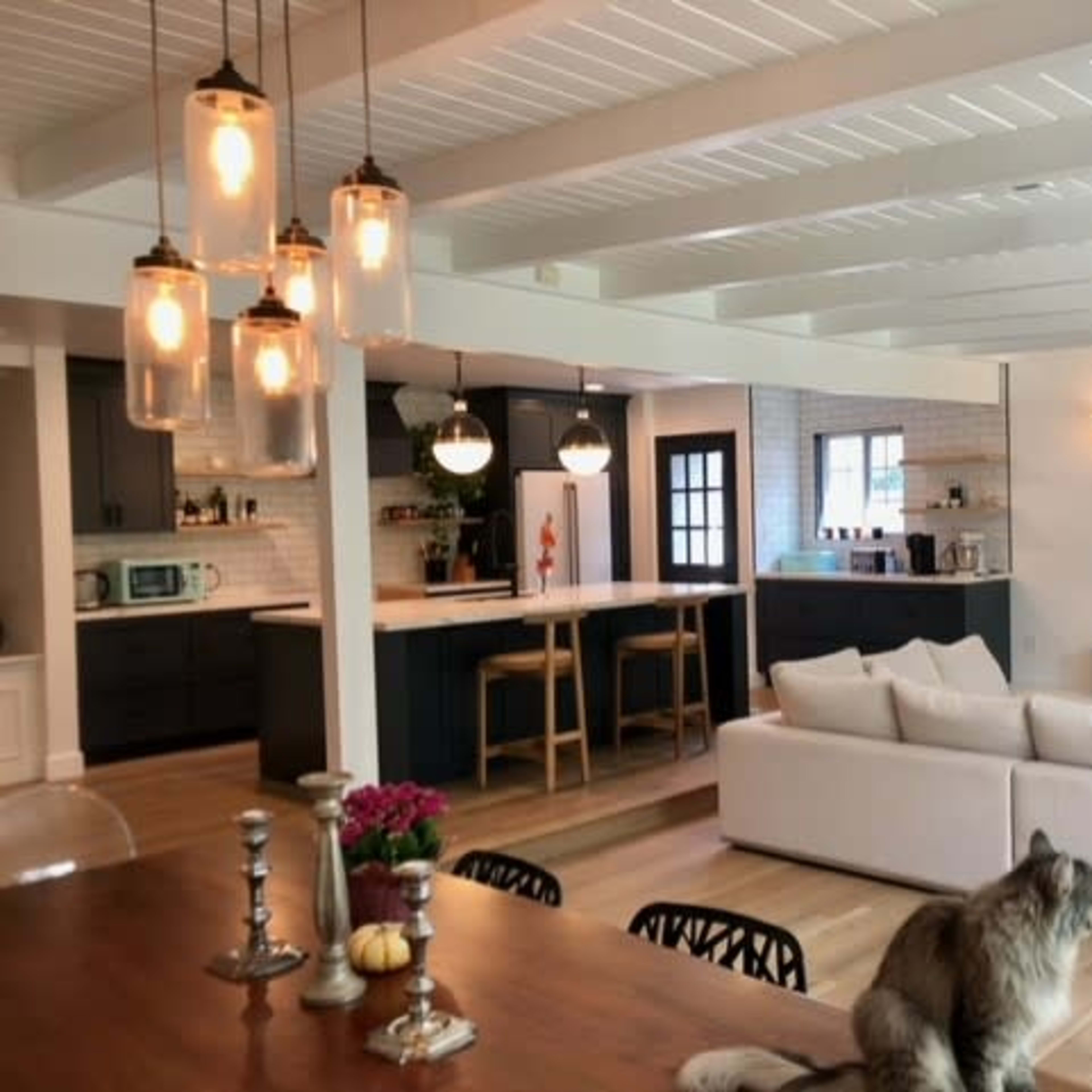 A modern open-concept living space with a dining area, kitchen, and living room, featuring white beams, pendant lights, and a pet on a couch.