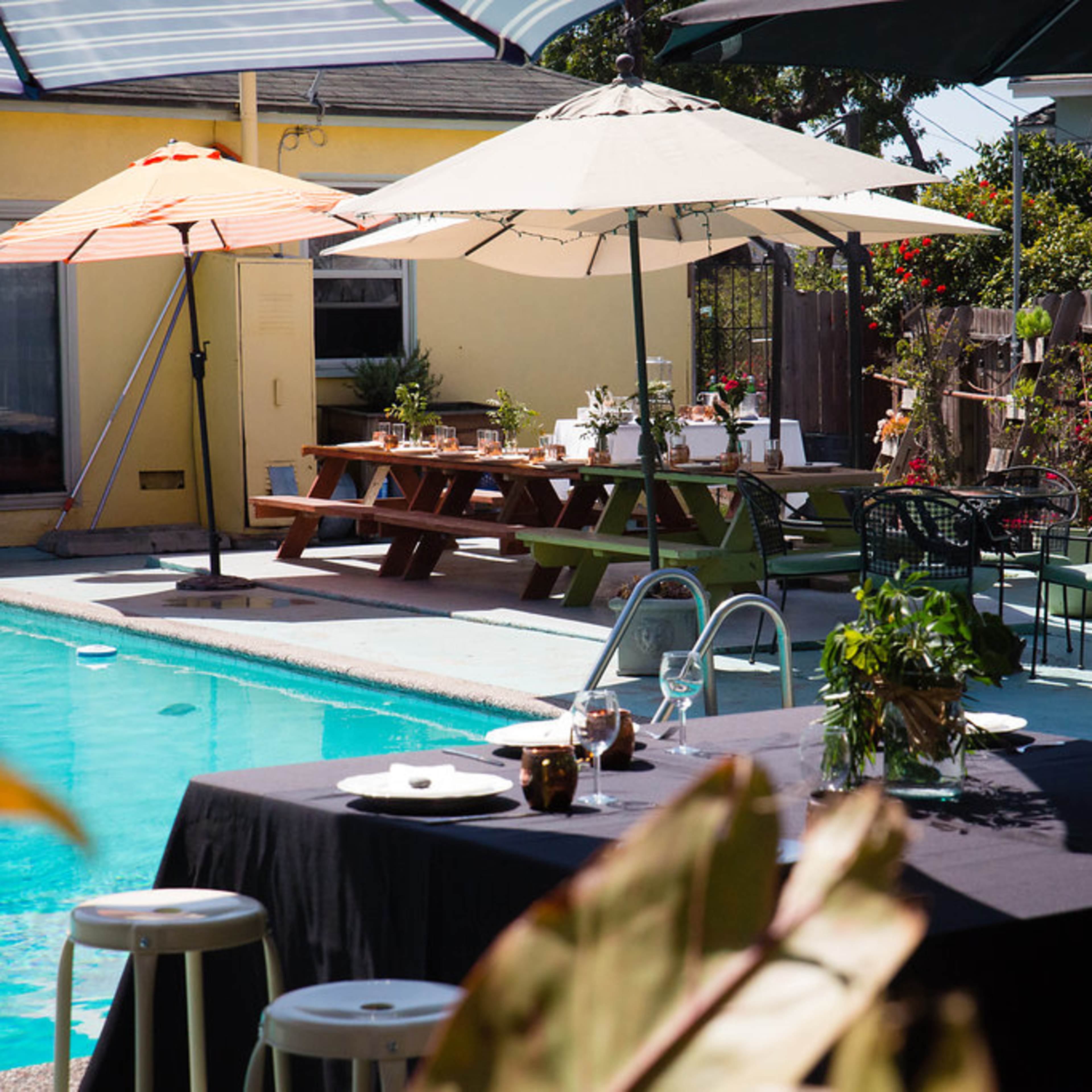 A backyard scene with a swimming pool, dining tables set for a meal, and various umbrellas providing shade.