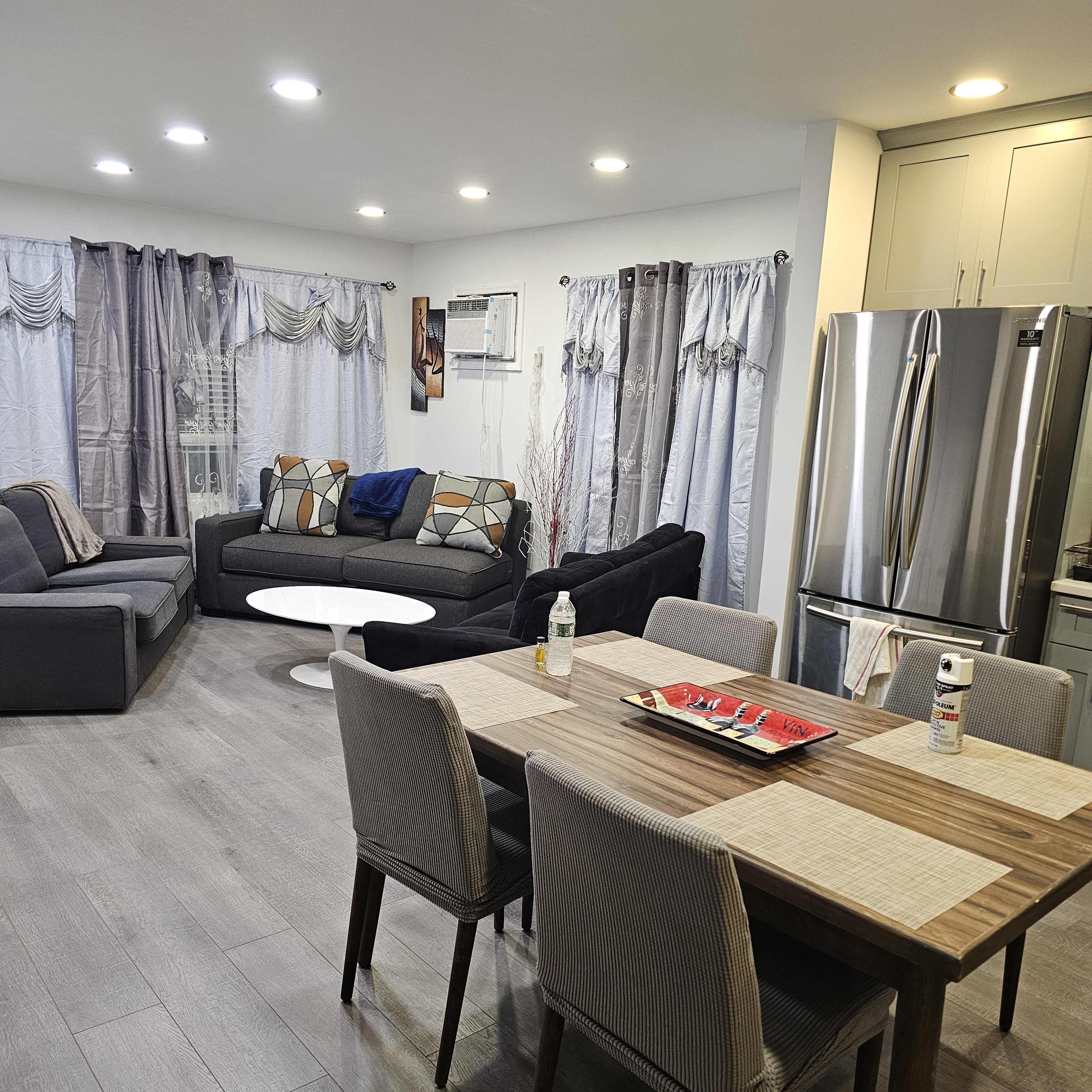 A modern living room and dining area with a gray sofa, a round coffee table, a dining table with chairs, and a stainless steel refrigerator in the kitchen area.