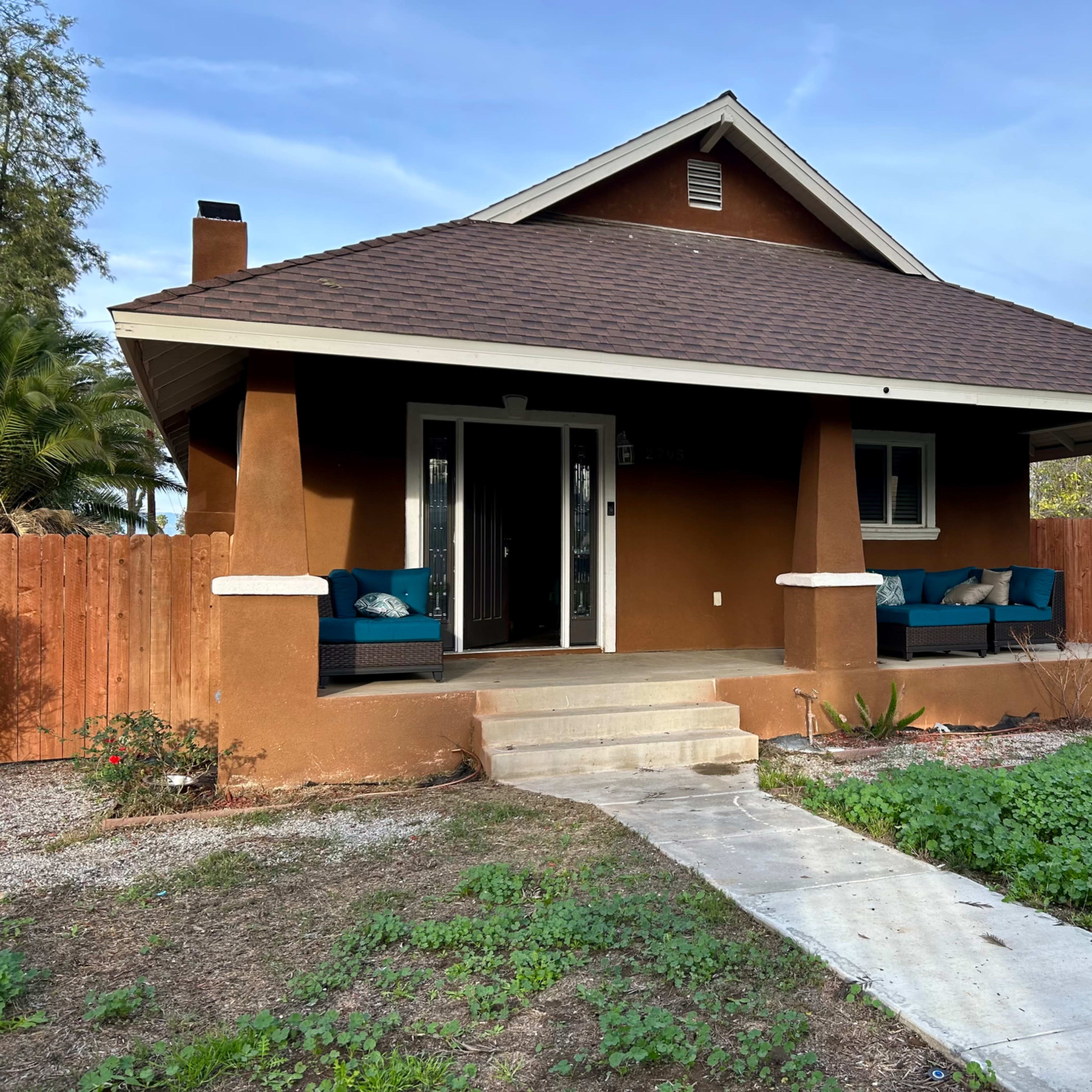 A brown bungalow with a front porch and two blue chairs sits in a yard with grass and gravel pathways.