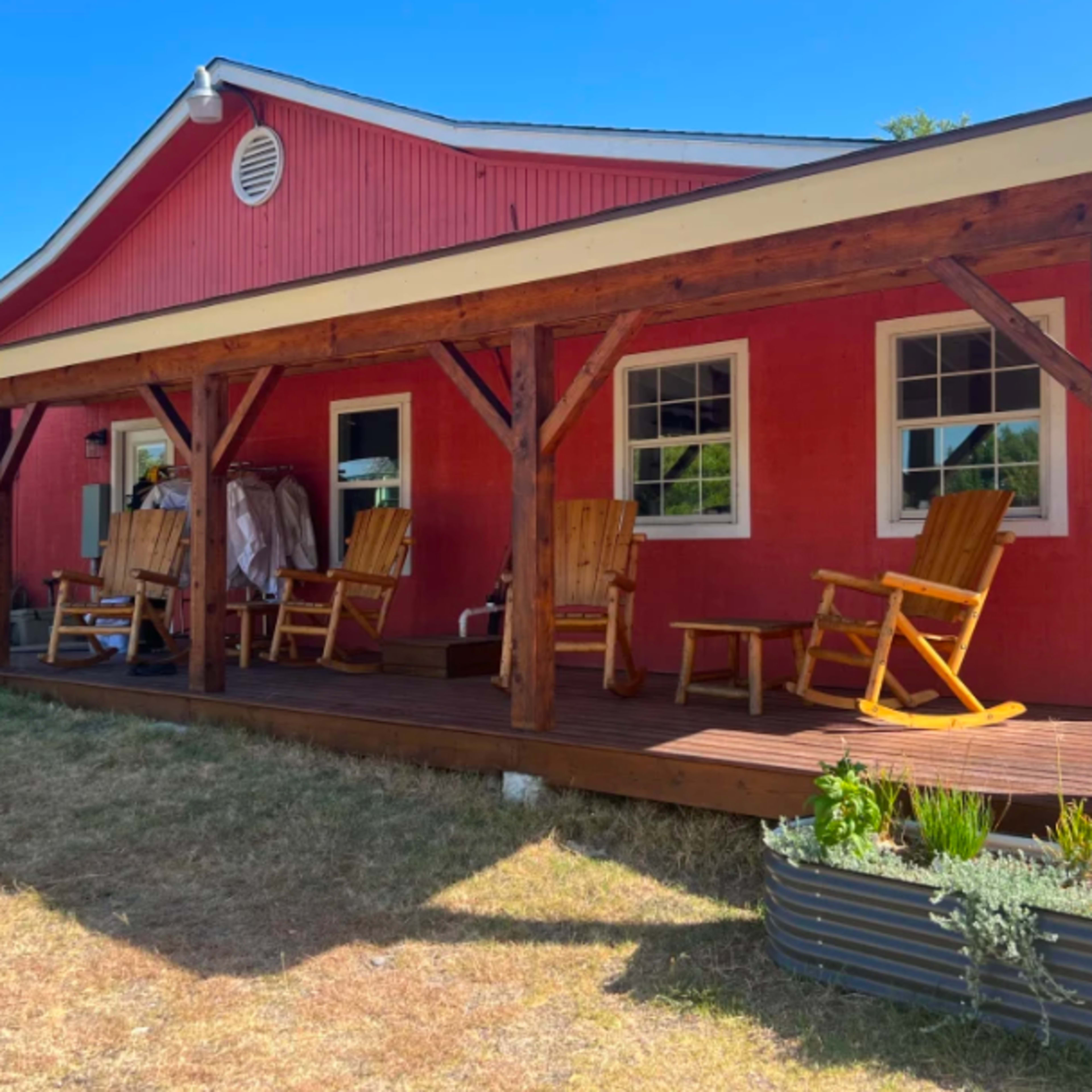 A red house with a wooden porch features several rocking chairs and plants along the side.