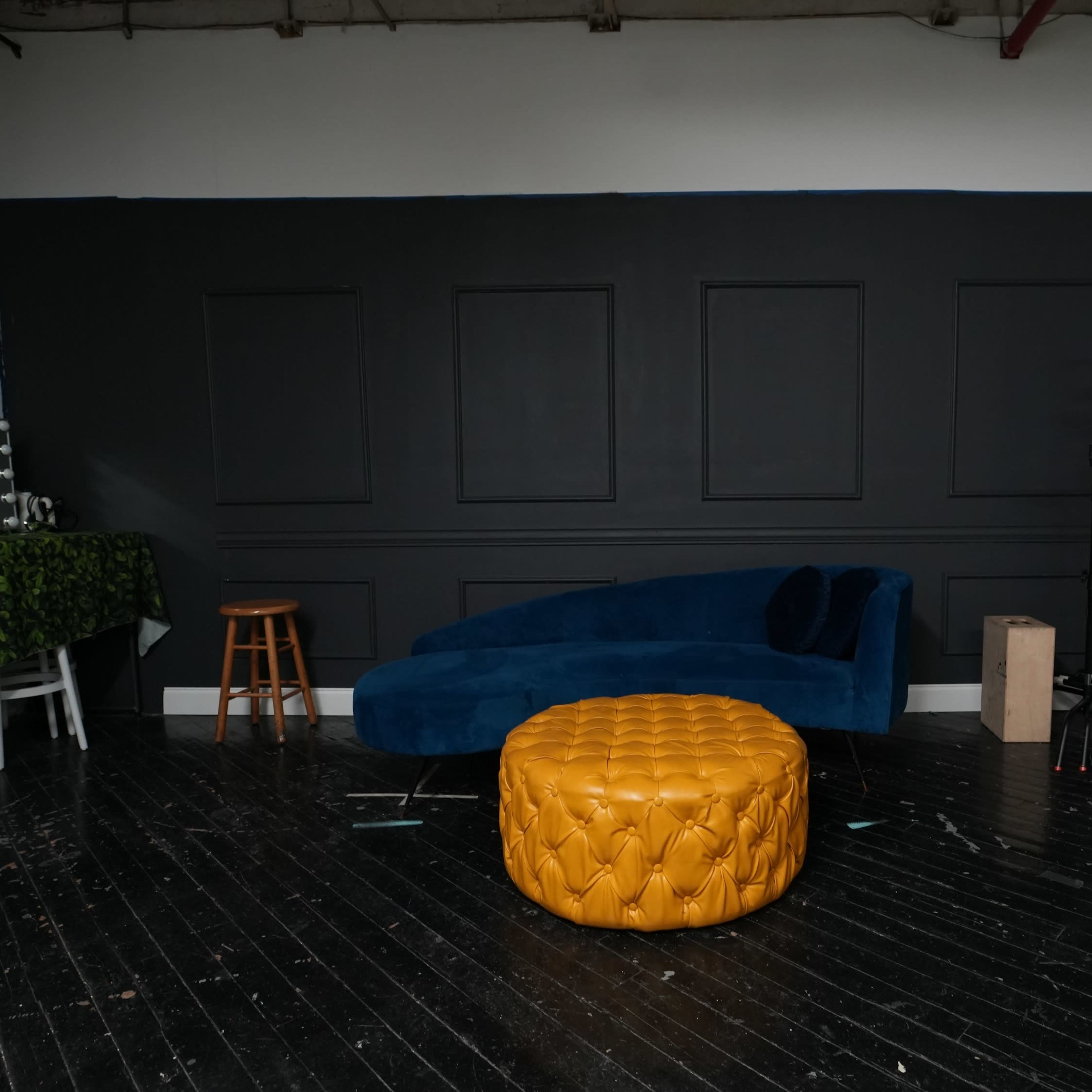 A spacious studio with a dark wall, a blue couch, a yellow ottoman, and various backdrops and props arranged around the room.