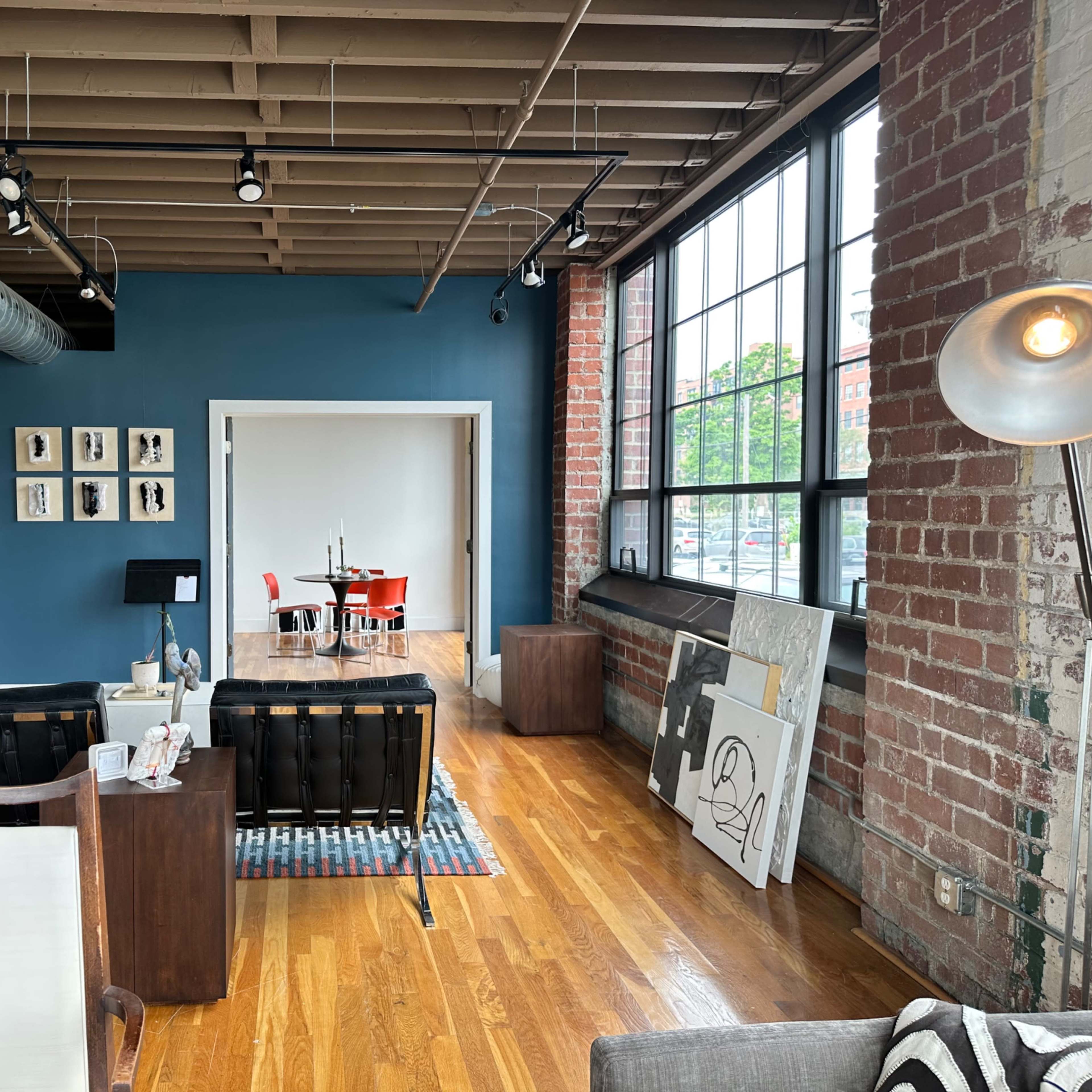 A spacious loft-style living area with hardwood floors, exposed brick walls, and modern furniture, including two black sofas and a round dining table in a separate room.