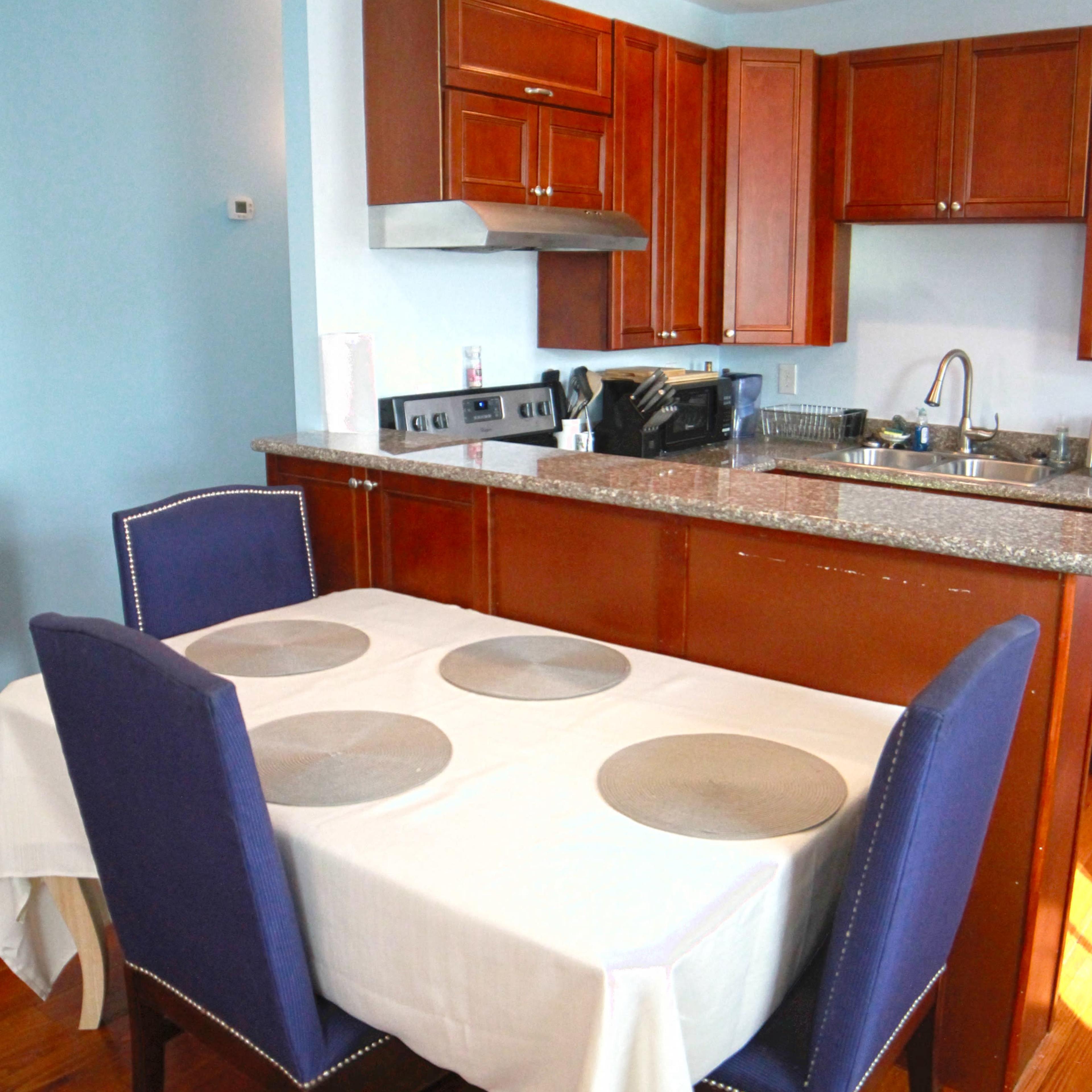 A kitchen area features a wooden table with four blue chairs, covered by a white tablecloth, beside cabinets and appliances.