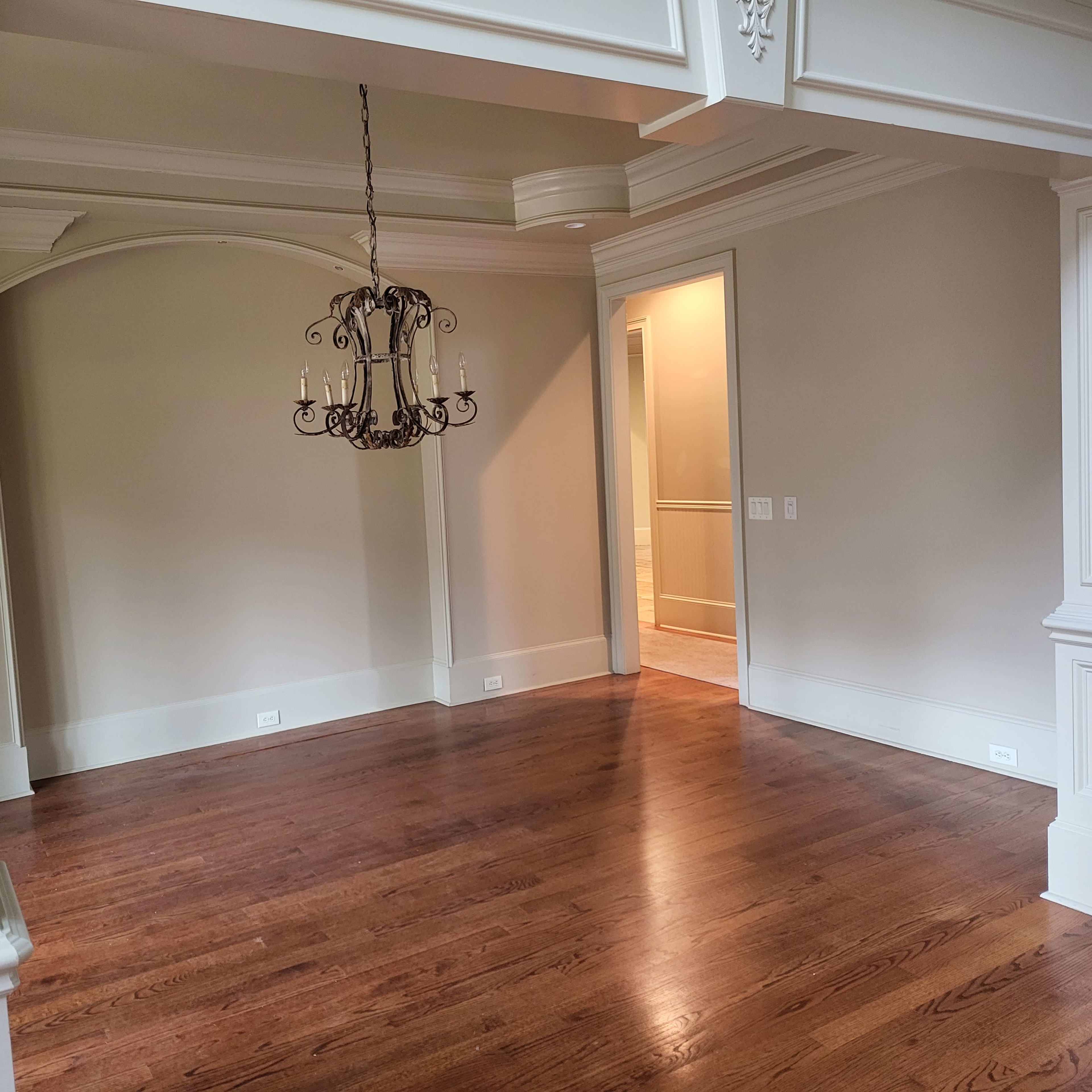 A spacious room with wooden flooring, featuring a chandelier and an archway leading to another area.