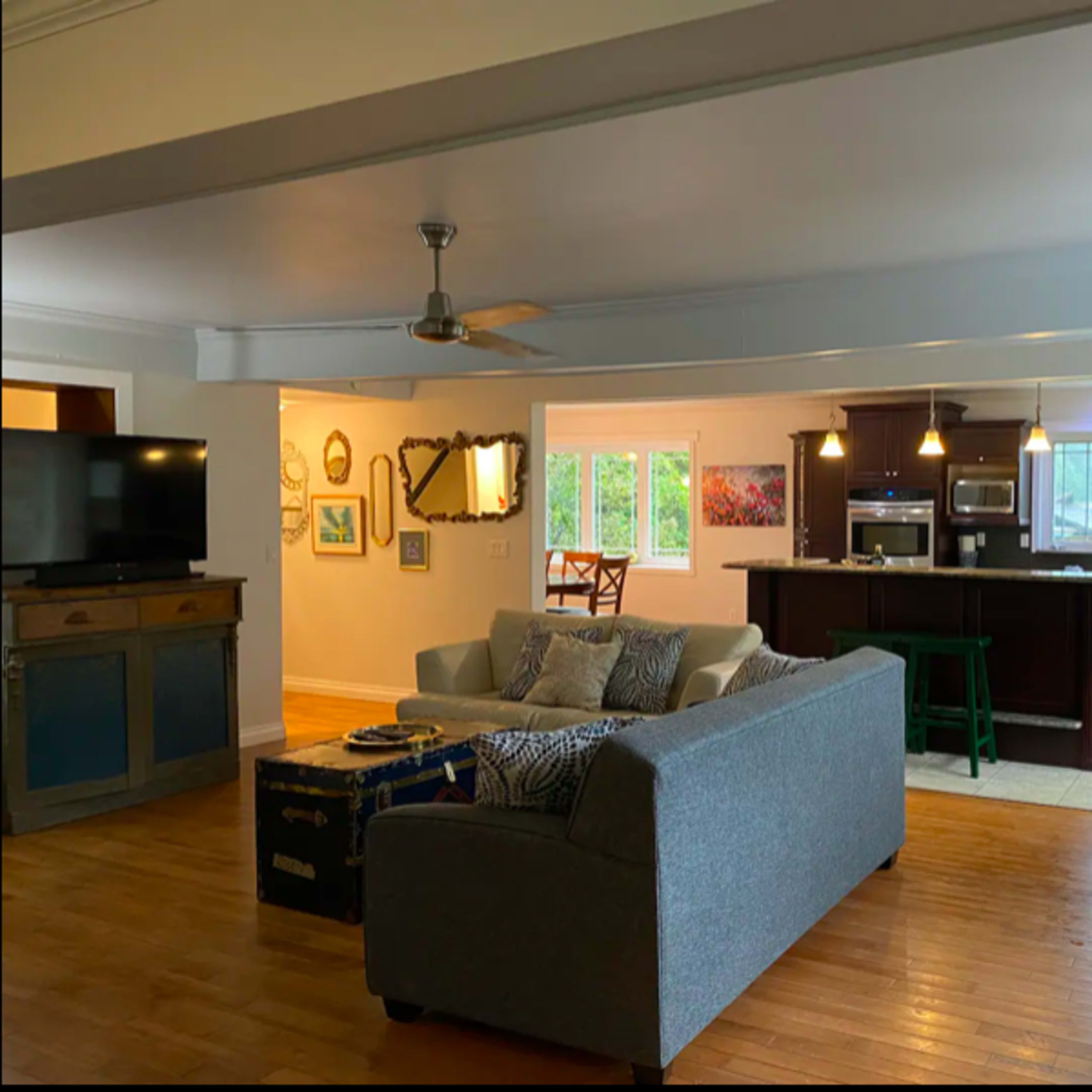 A spacious living area with a gray sofa, a dining area with wooden chairs, and a kitchen featuring dark cabinetry and stainless steel appliances.