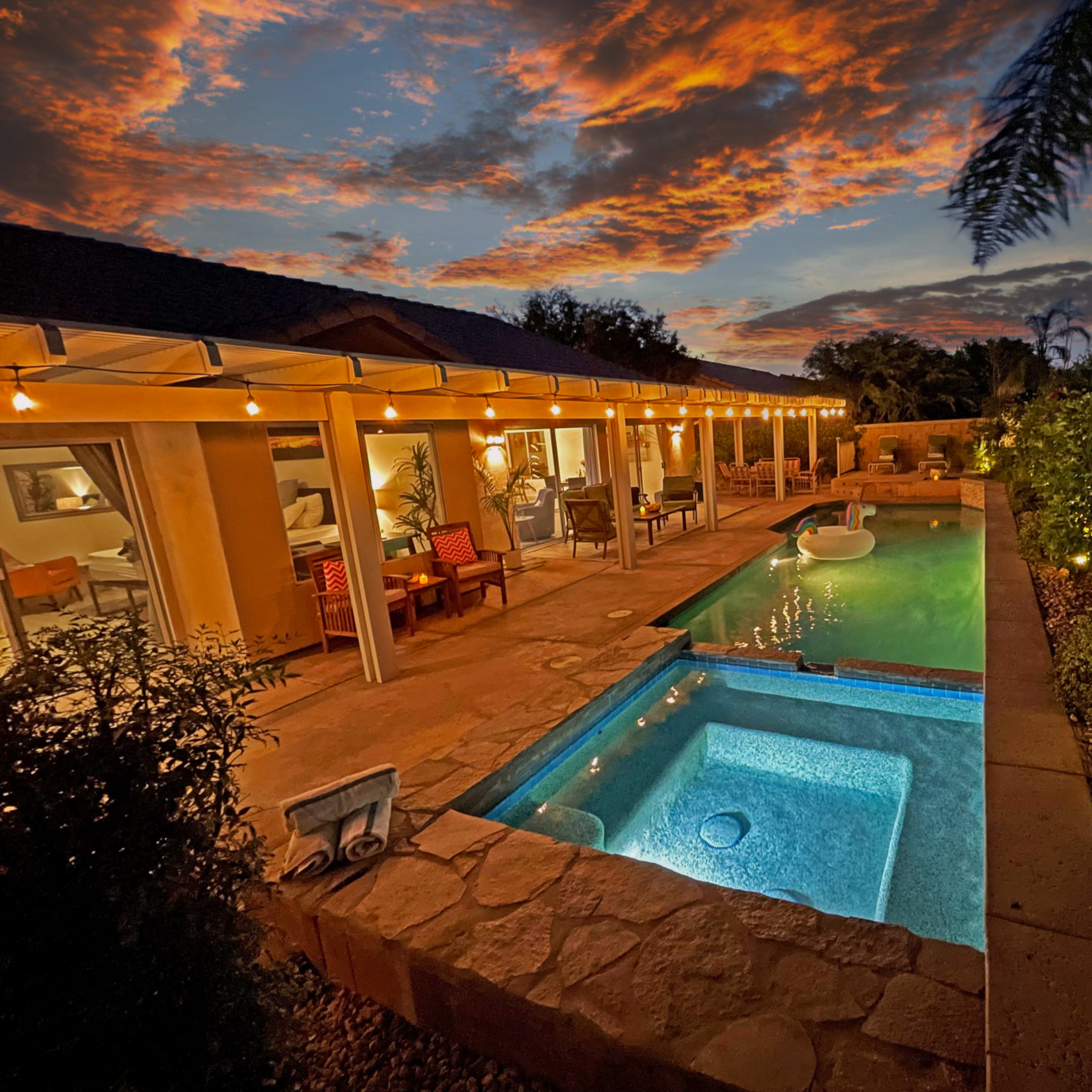 A well-lit patio with a swimming pool and spa is set against a vibrant sunset sky.