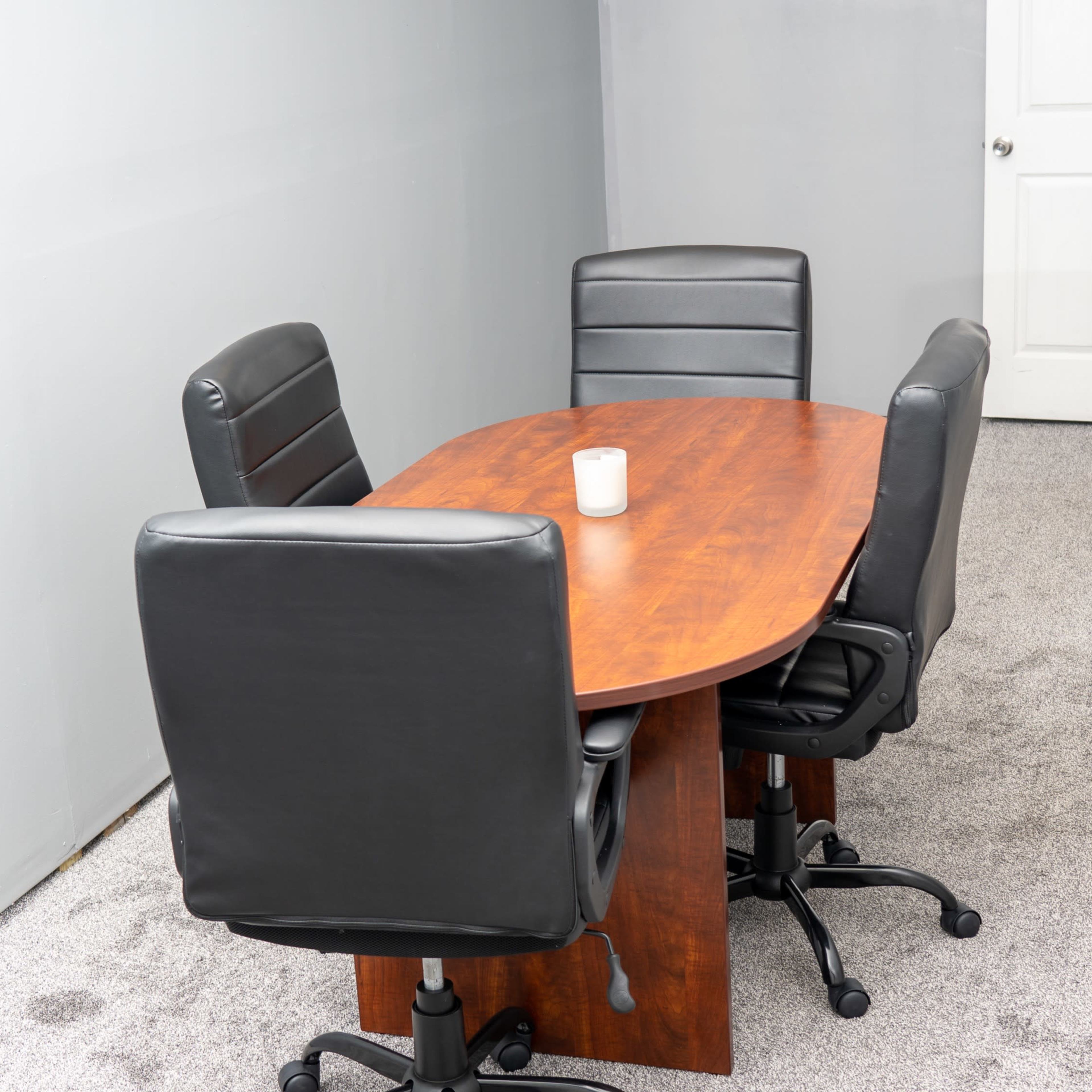 A wooden oval table is surrounded by four black office chairs in a carpeted meeting room with gray walls.