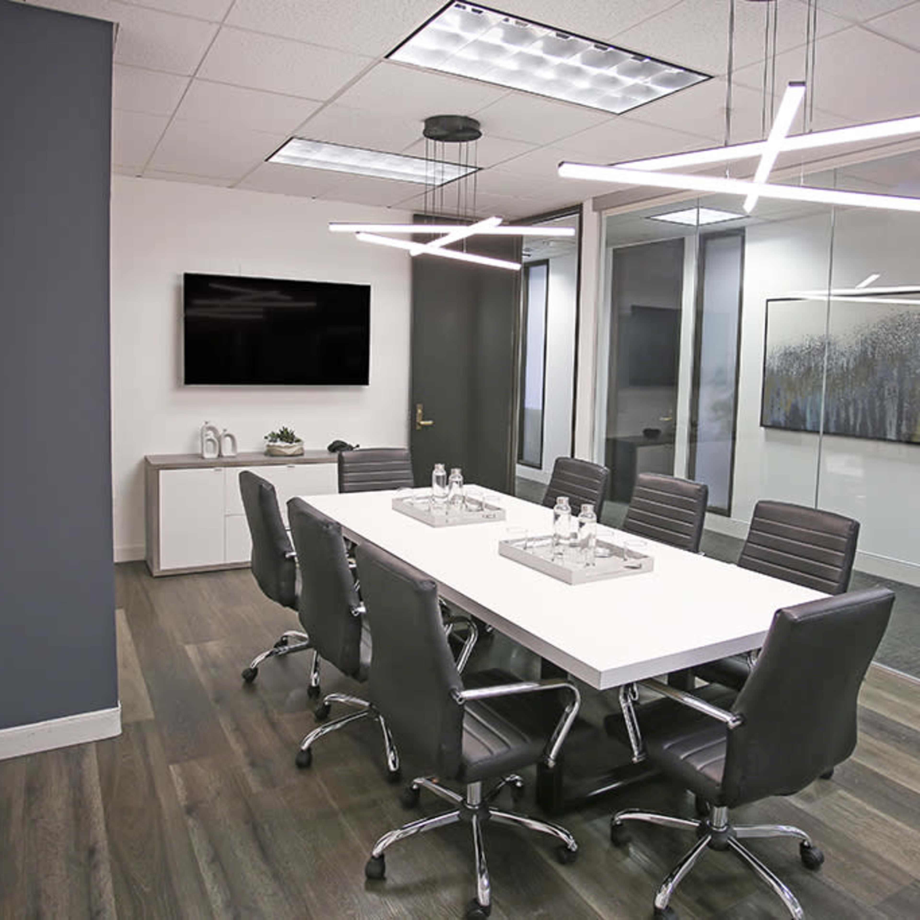 A modern conference room featuring a long white table surrounded by black chairs, with a television mounted on the wall and a glass partition.