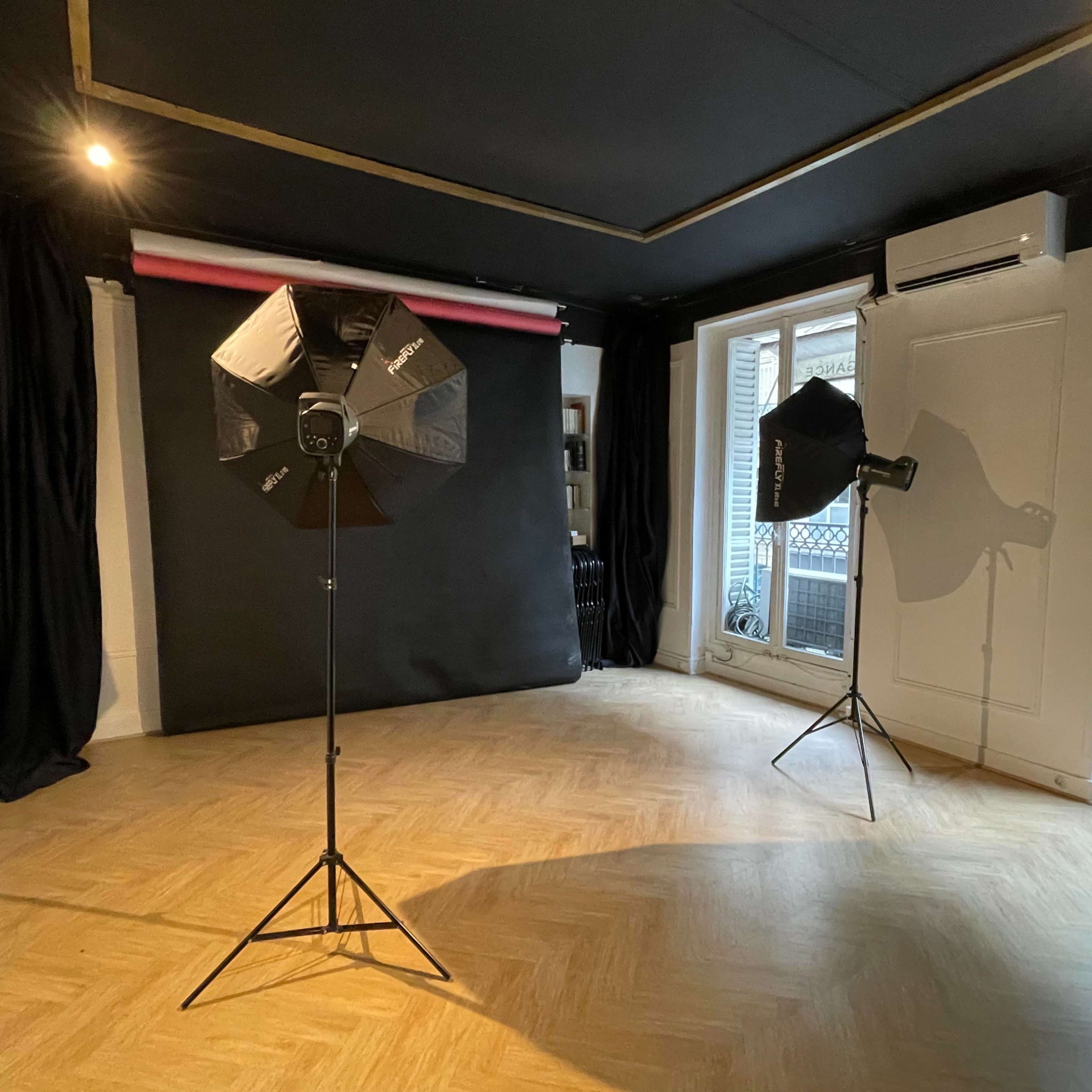 A photo studio featuring two softbox light stands, a black backdrop, and large windows with balcony views.