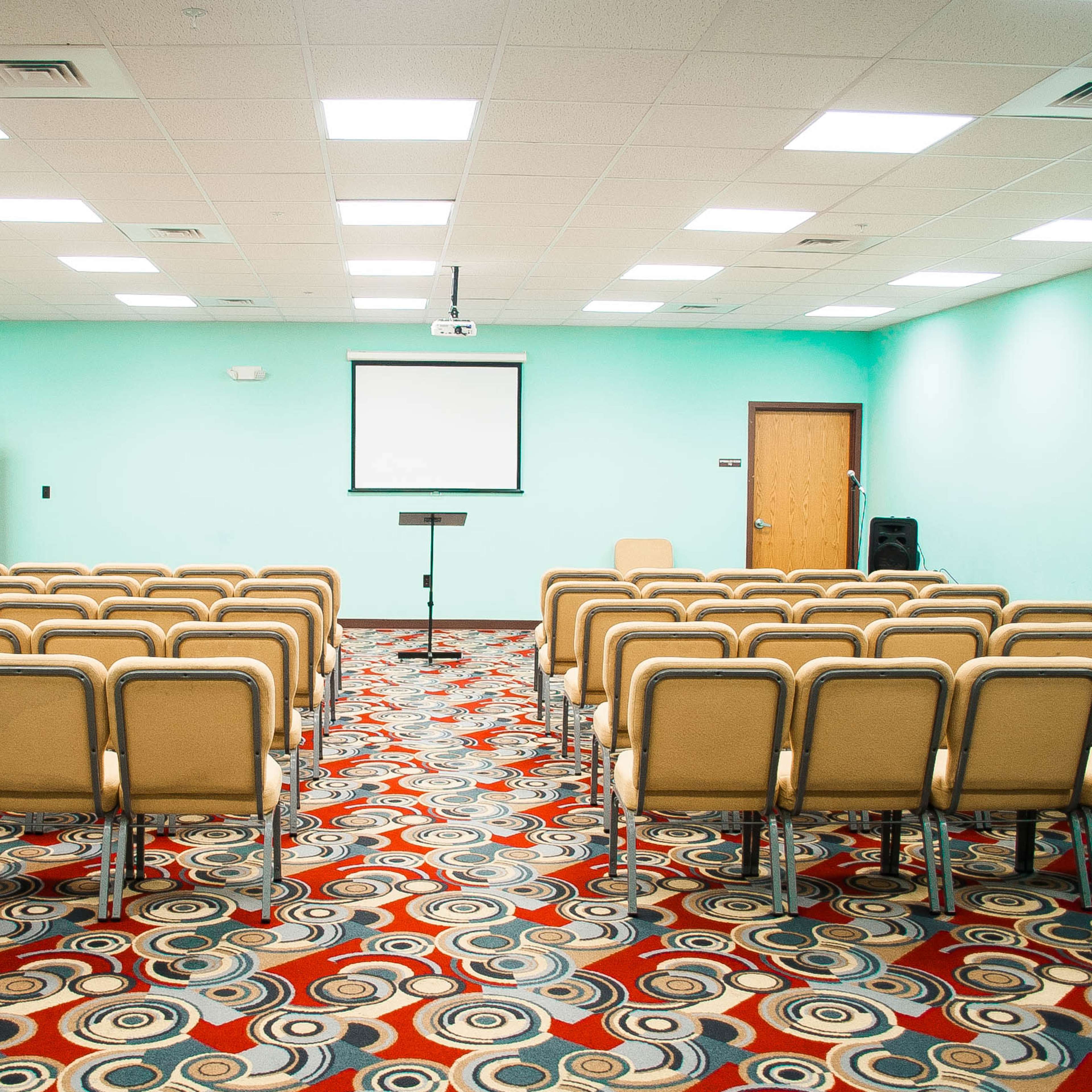 A meeting room with rows of beige chairs facing a screen, set against a light blue wall and patterned carpet.