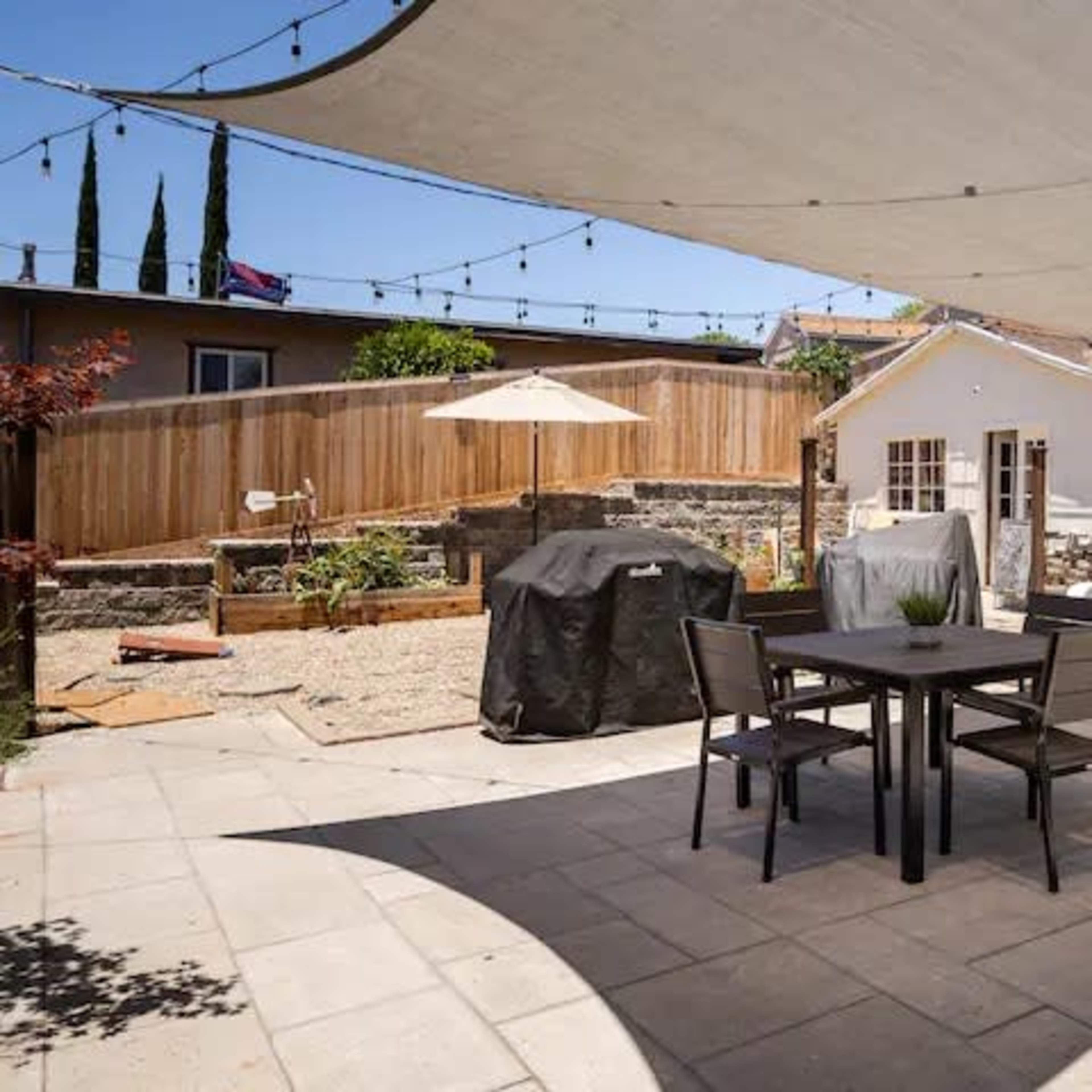 A patio area features a rectangular table and chairs under a large shaded canopy, with a barbecue grill covered nearby and garden features in the background.